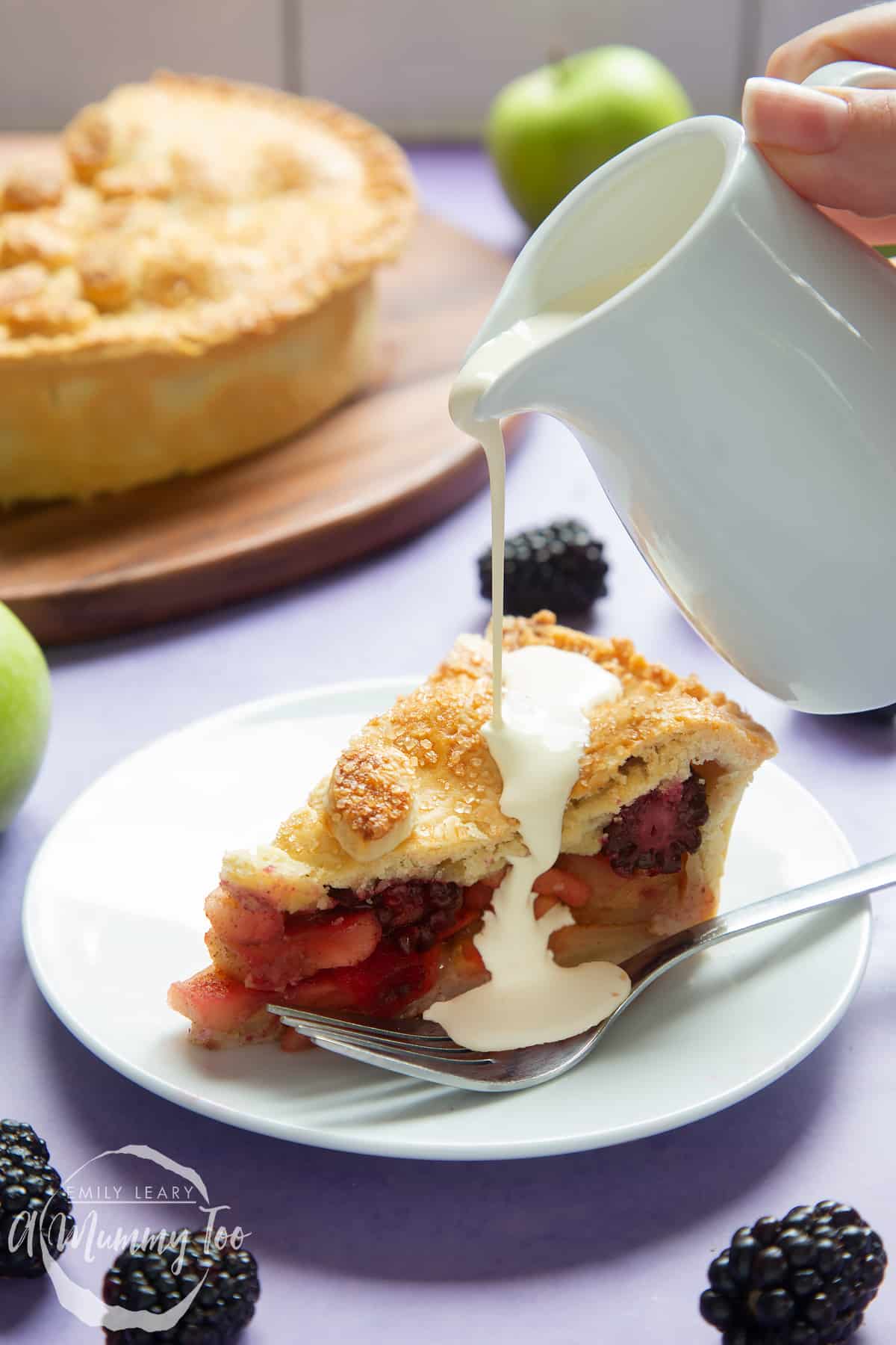 A slice of apple and blackberry pie on a white plate with a fork. A small white jug pours cream onto the pie.