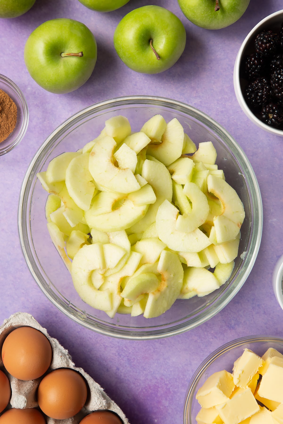 Apple slices in a glass mixing bowl. Ingredients to make apple and blackberry pie surround the bowl.