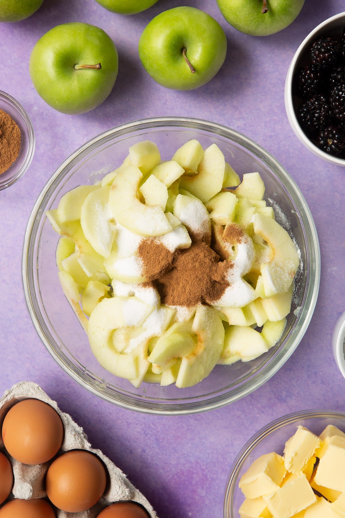Apple slices in a glass mixing bowl with sugar and cinnamon. Ingredients to make apple and blackberry pie surround the bowl.