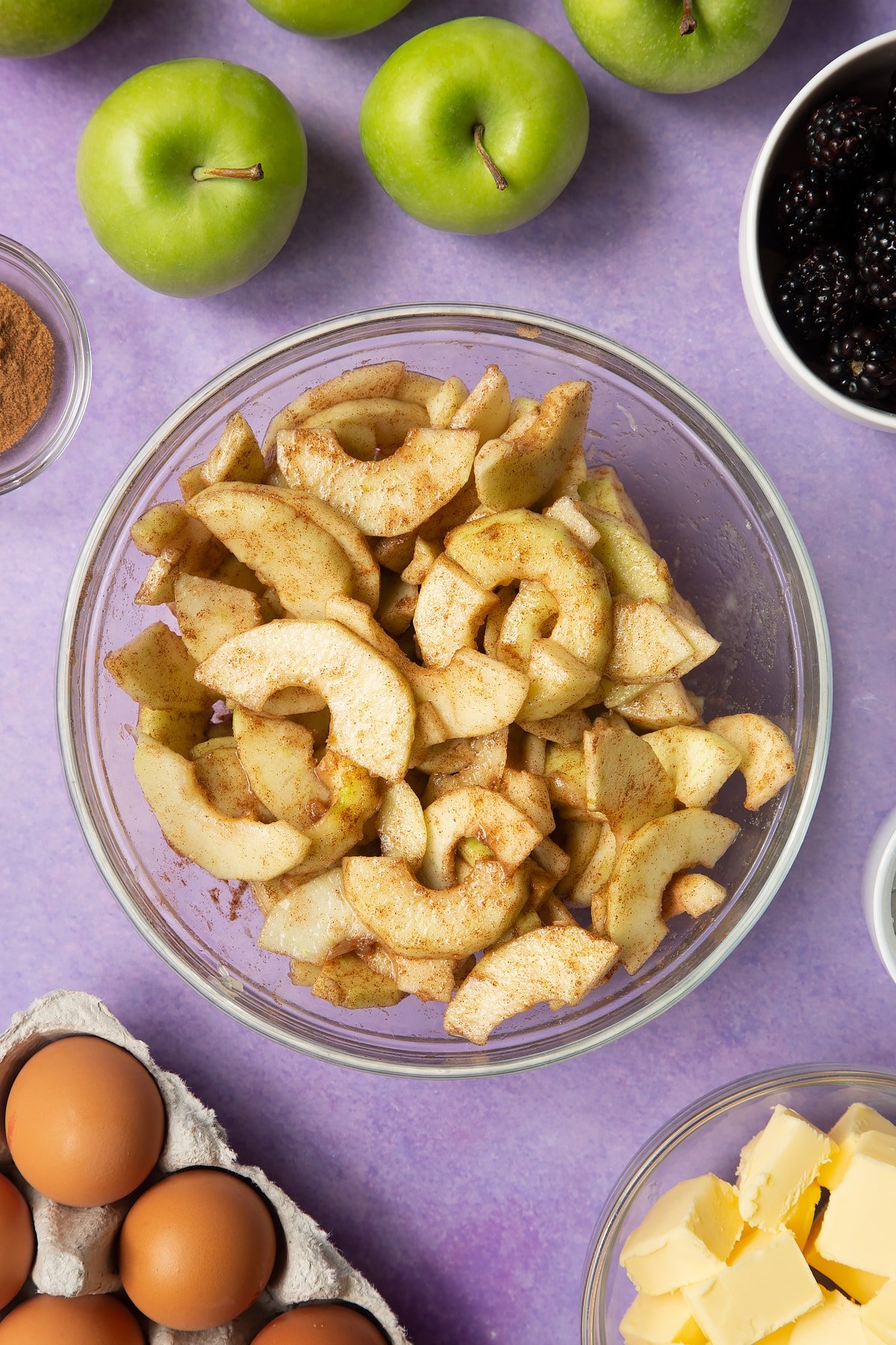 Apple slices mixed with with sugar and cinnamon in a glass mixing bowl. Ingredients to make apple and blackberry pie surround the bowl.