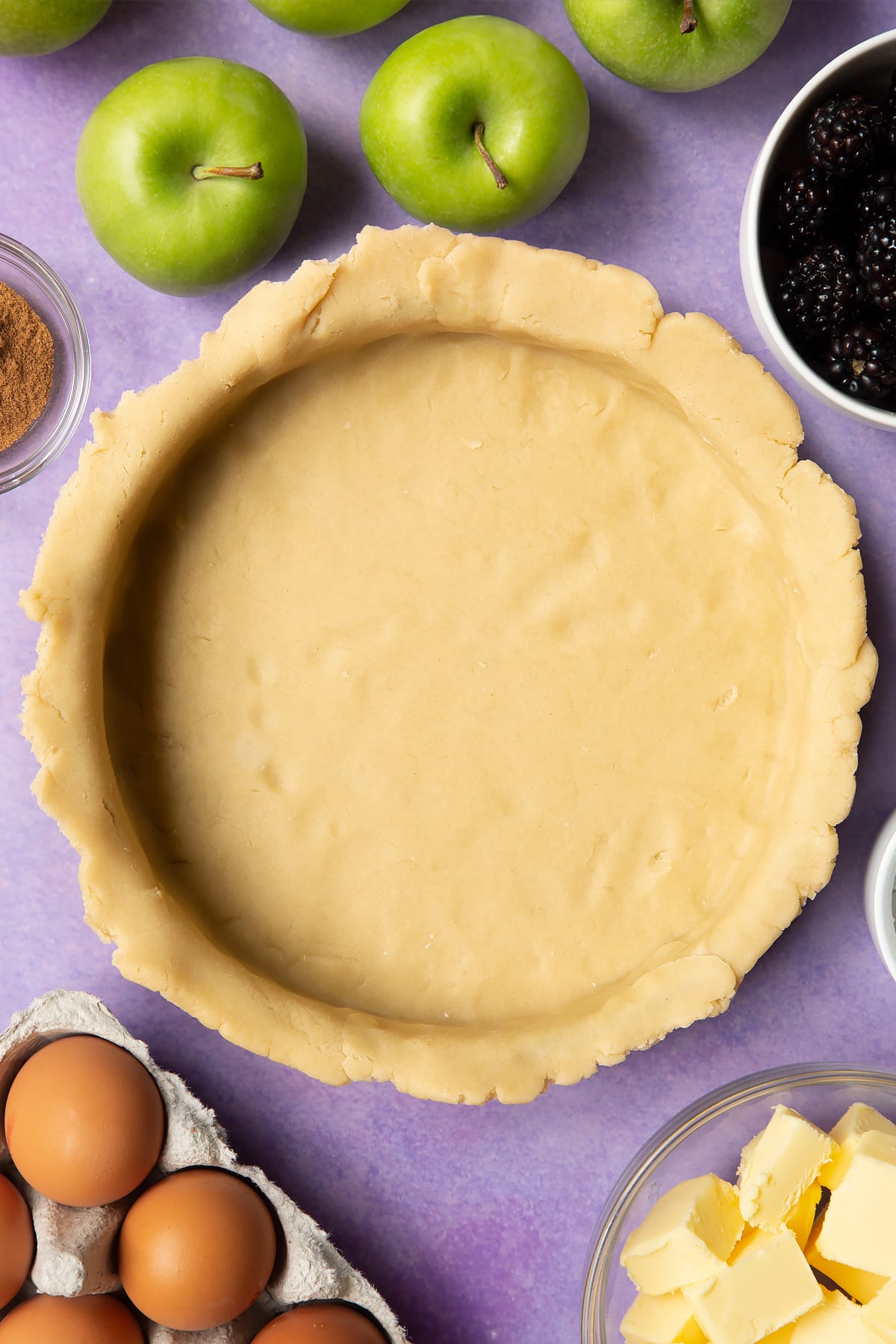 A pie tin lined with pastry. Ingredients to make apple and blackberry pie surround the tin.