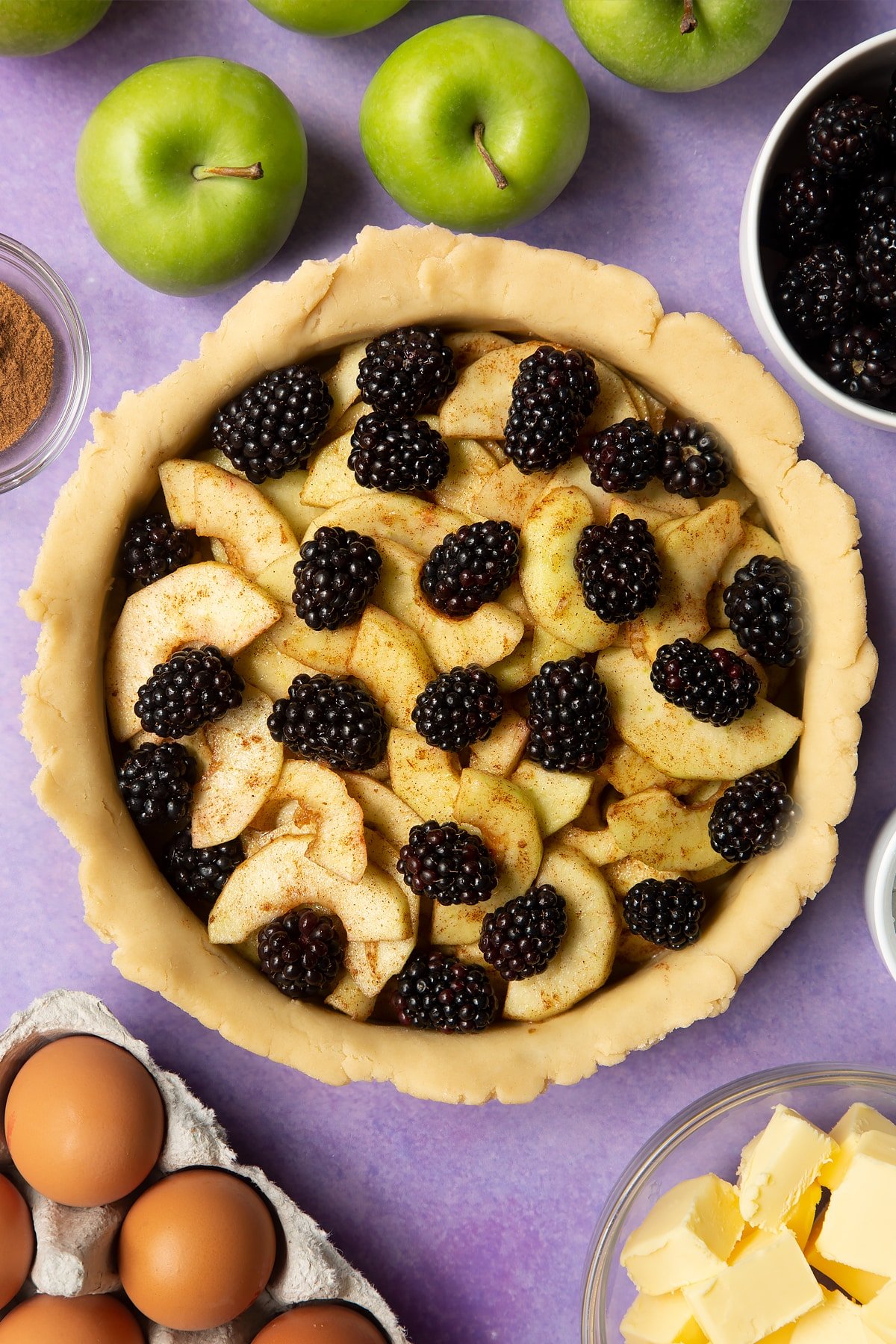 A pie tin lined with pastry and filled with apple slices and blackberries. Ingredients to make apple and blackberry pie surround the tin.