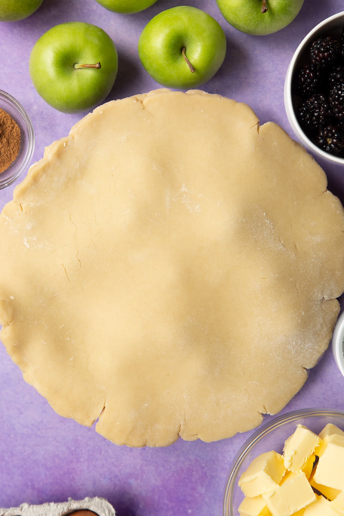 A pie tin lined with pastry and filled with apple slices and blackberries, with another disc of pastry resting on top. Ingredients to make apple and blackberry pie surround the tin.