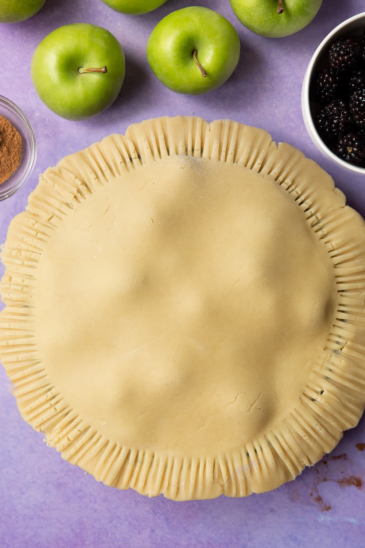 A pastry pie filled with apple slices and blackberries, a fork has been pressed around the edge. Ingredients to make apple and blackberry pie surround the tin.