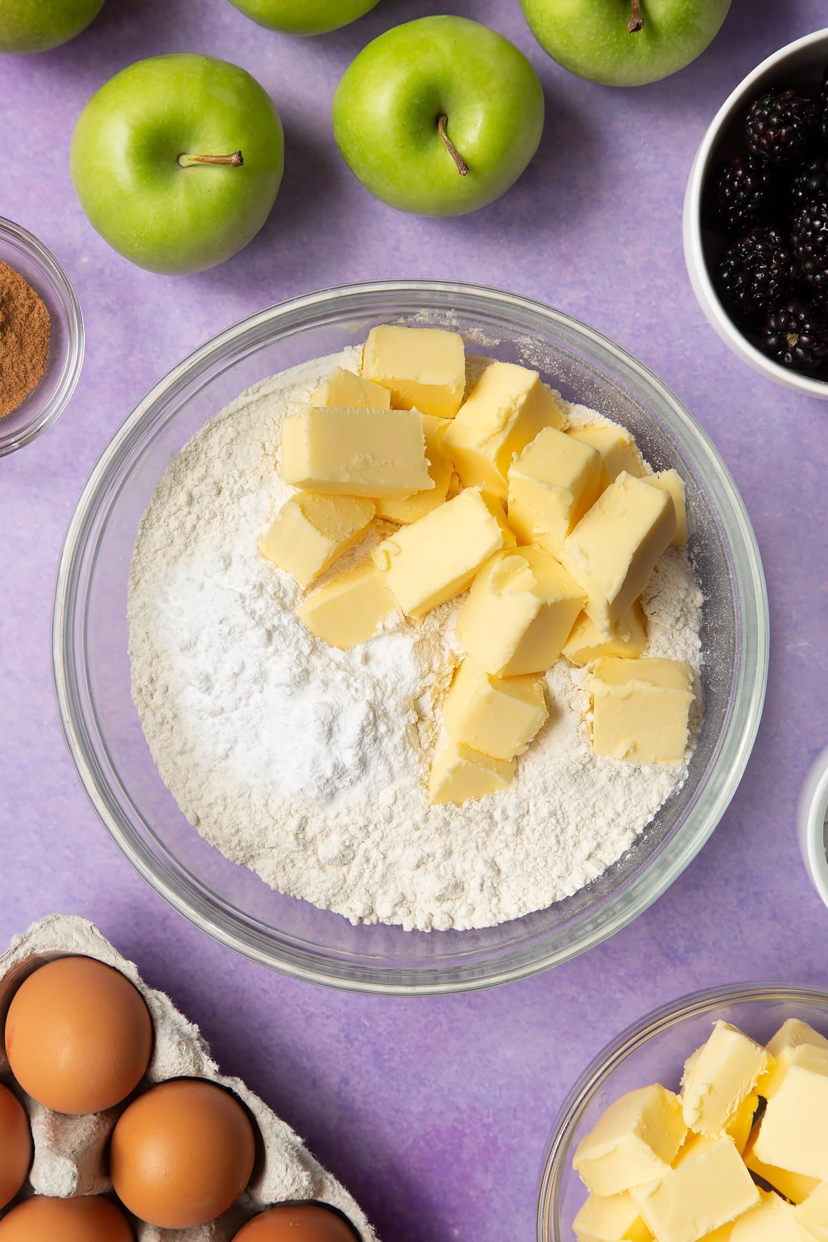 Flour, icing sugar and butter in a glass mixing bowl. Ingredients to make apple and blackberry pie surround the bowl.