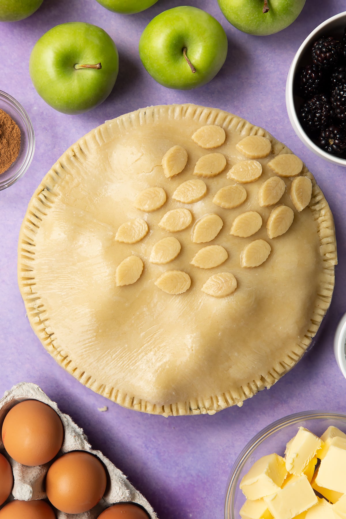 A raw apple and blackberry pie in a tin. The top has been brushed with egg white and decorated with pastry leaves.