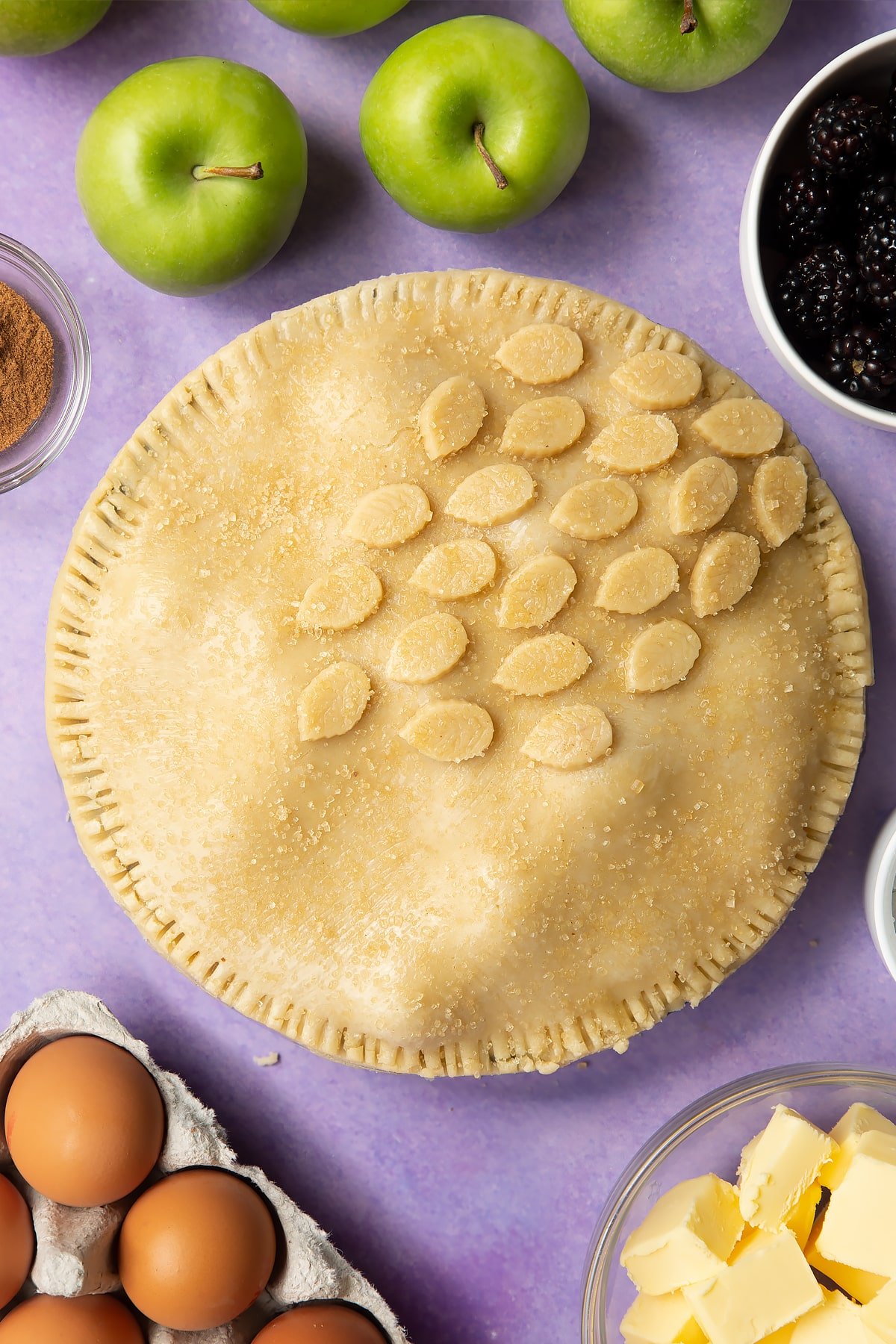 A raw apple and blackberry pie in a tin. The top has been brushed with egg white, decorated with pastry leaves and sprinkled with sugar.