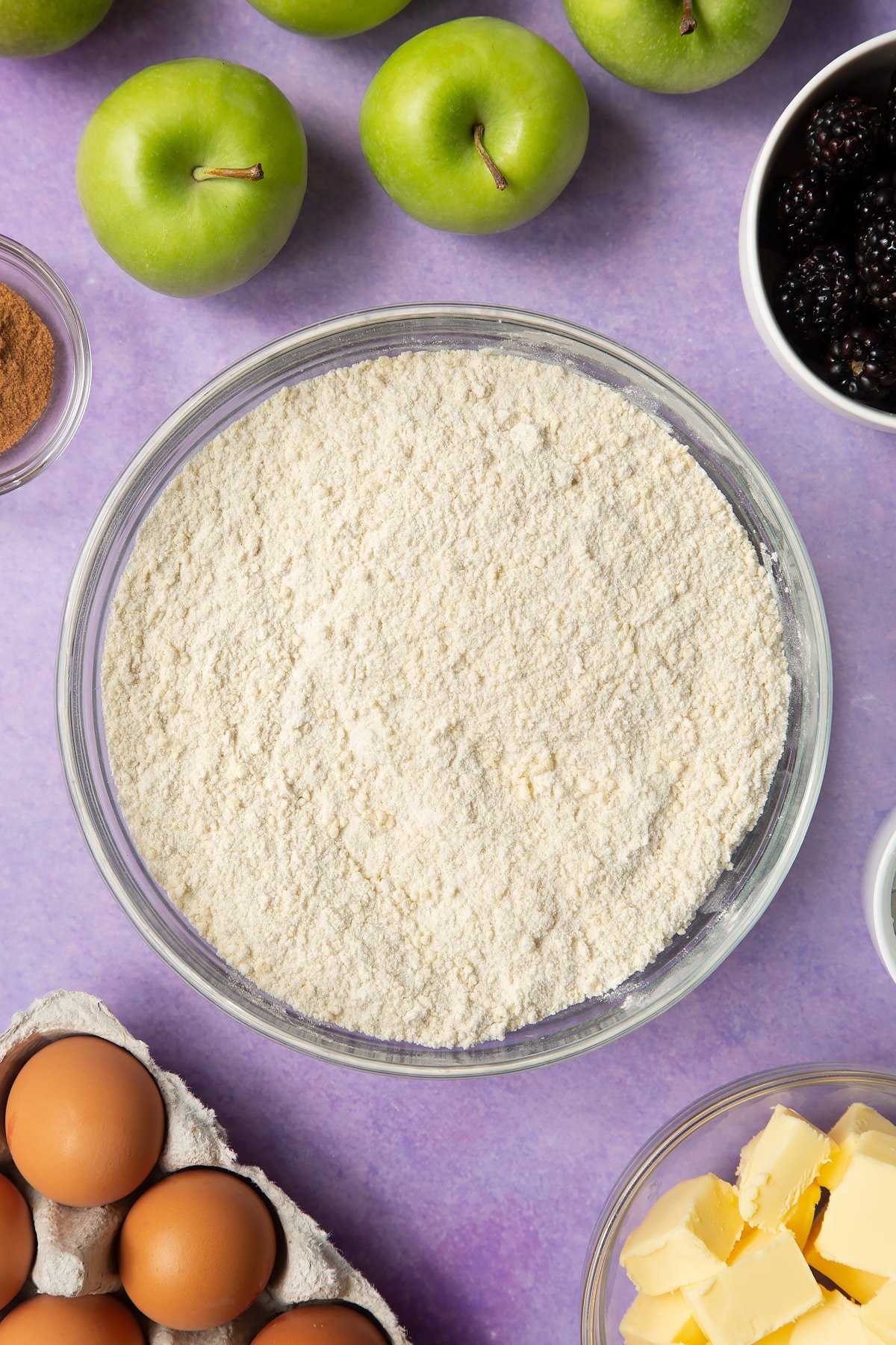 Flour, icing sugar and butter combined to a crumb in a glass mixing bowl. Ingredients to make apple and blackberry pie surround the bowl.