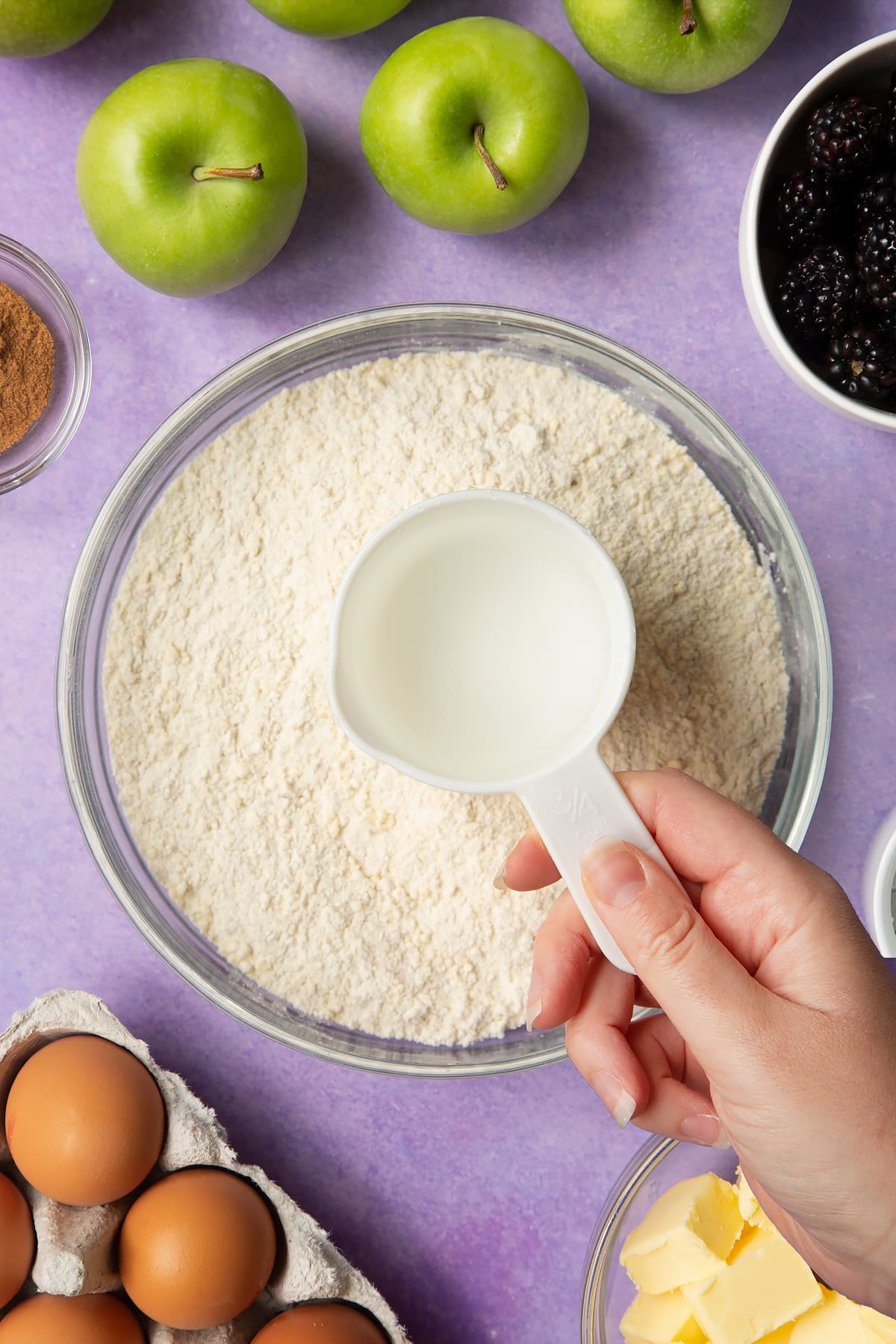 A flour and butter in a glass mixing bowl. A hand holds a cup of water above the bowl. Ingredients to make apple and blackberry pie surround the bowl.
