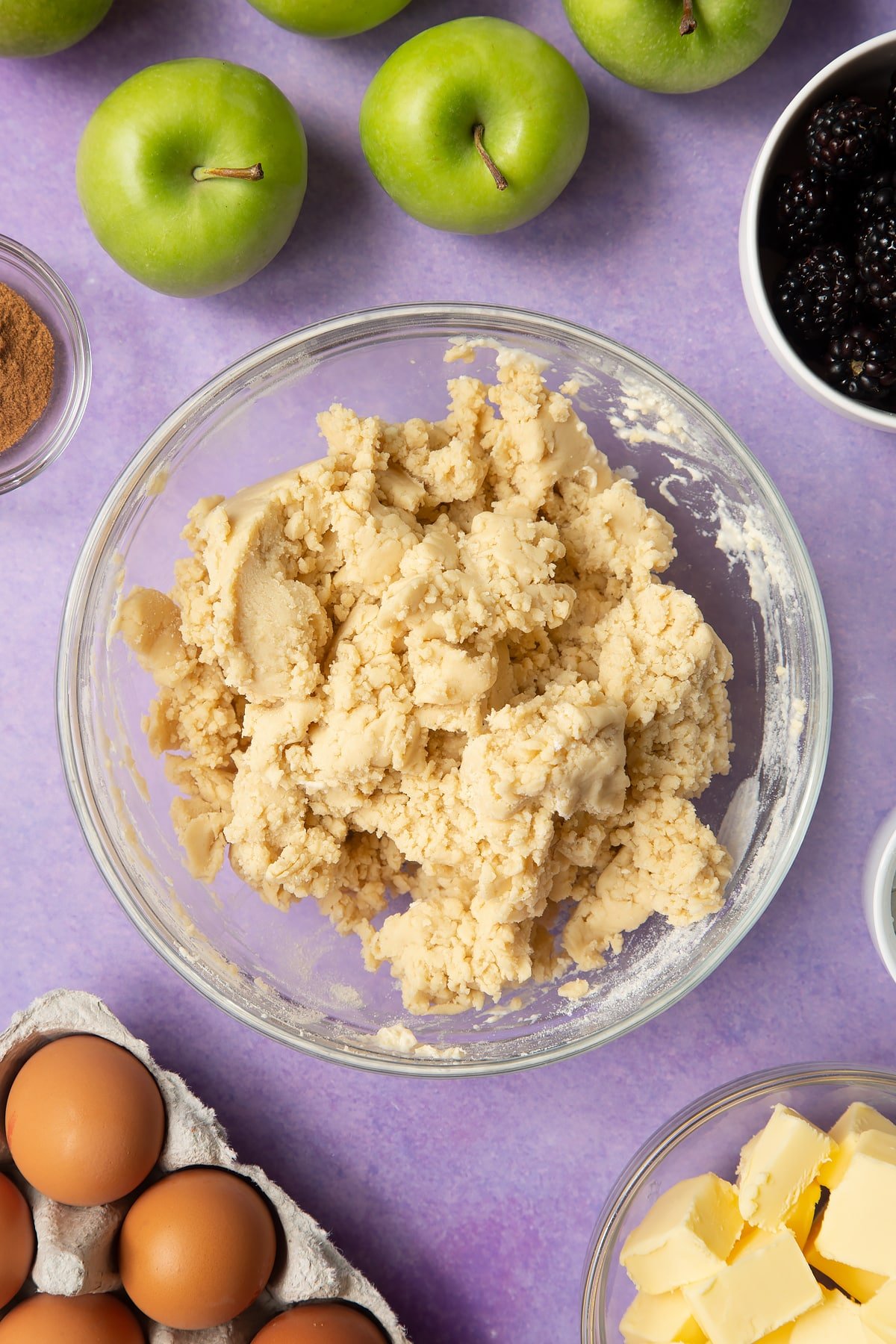 Pastry mix in a glass mixing bowl. Ingredients to make apple and blackberry pie surround the bowl.