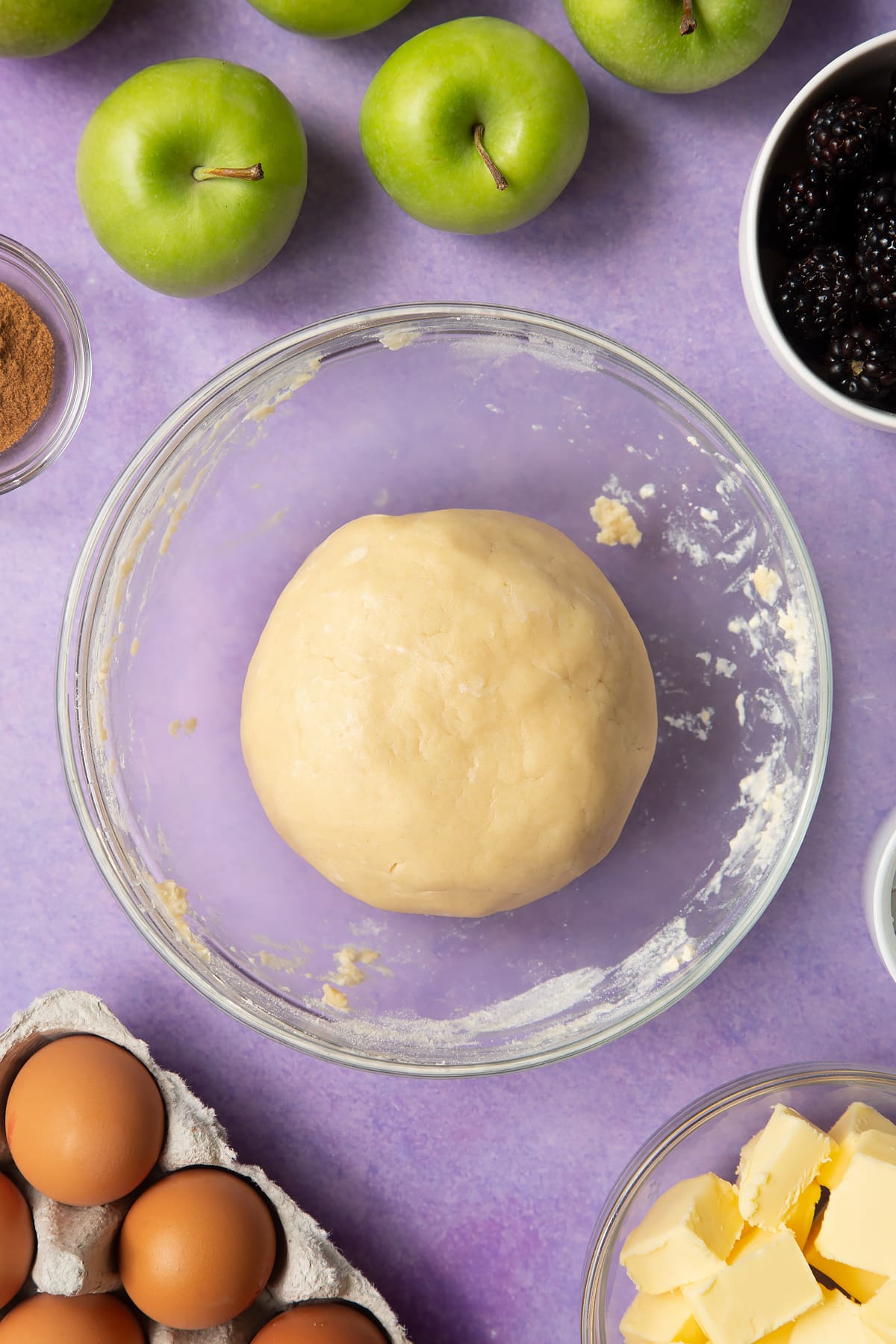 A ball of pastry in a glass mixing bowl. Ingredients to make apple and blackberry pie surround the bowl.