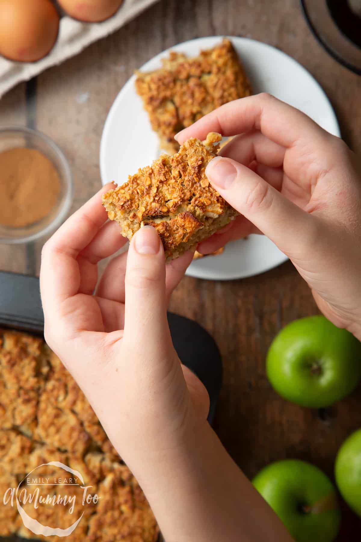 Hands holding a porridge square. Apples and more of the porridge squares are shown in the background.