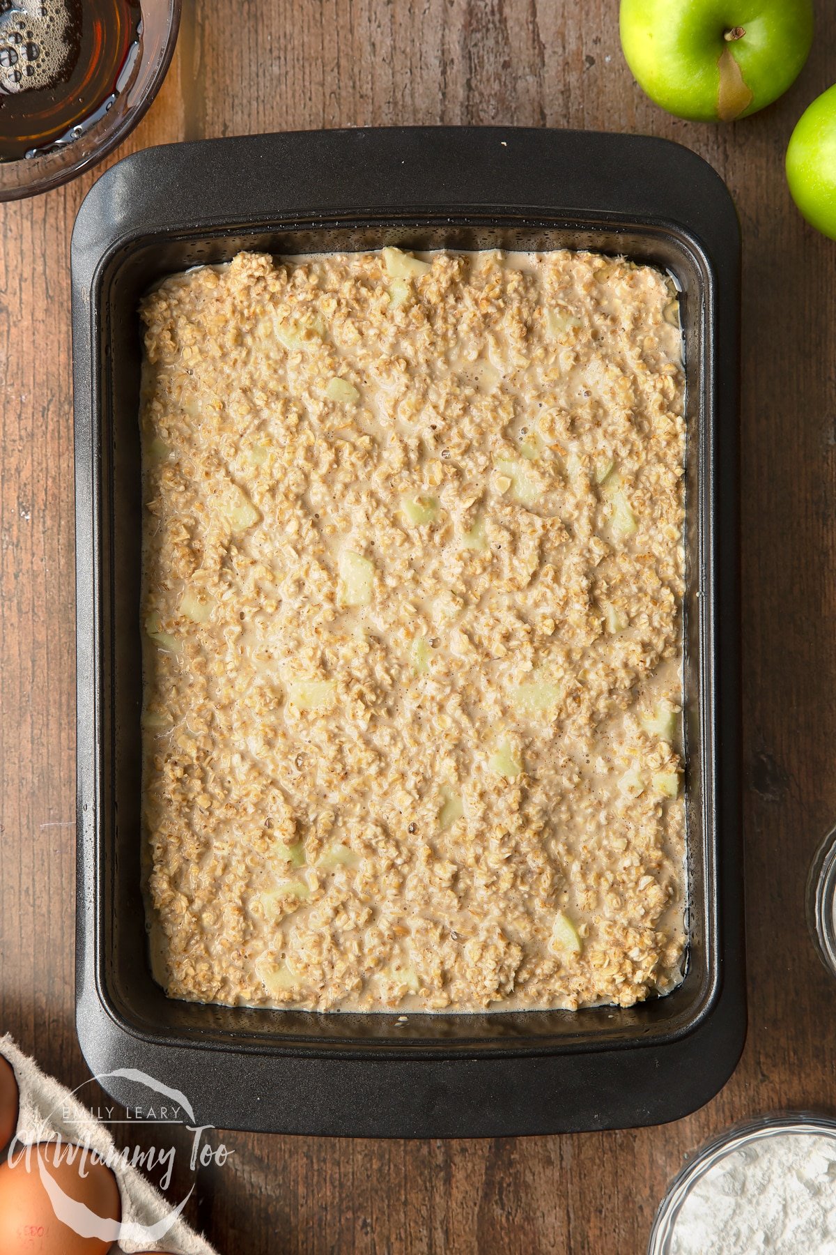 Oats, baking powder, salt, milk, eggs, maple syrup and apple mixed together in a tray. Ingredients to make porridge squares surround the bowl.