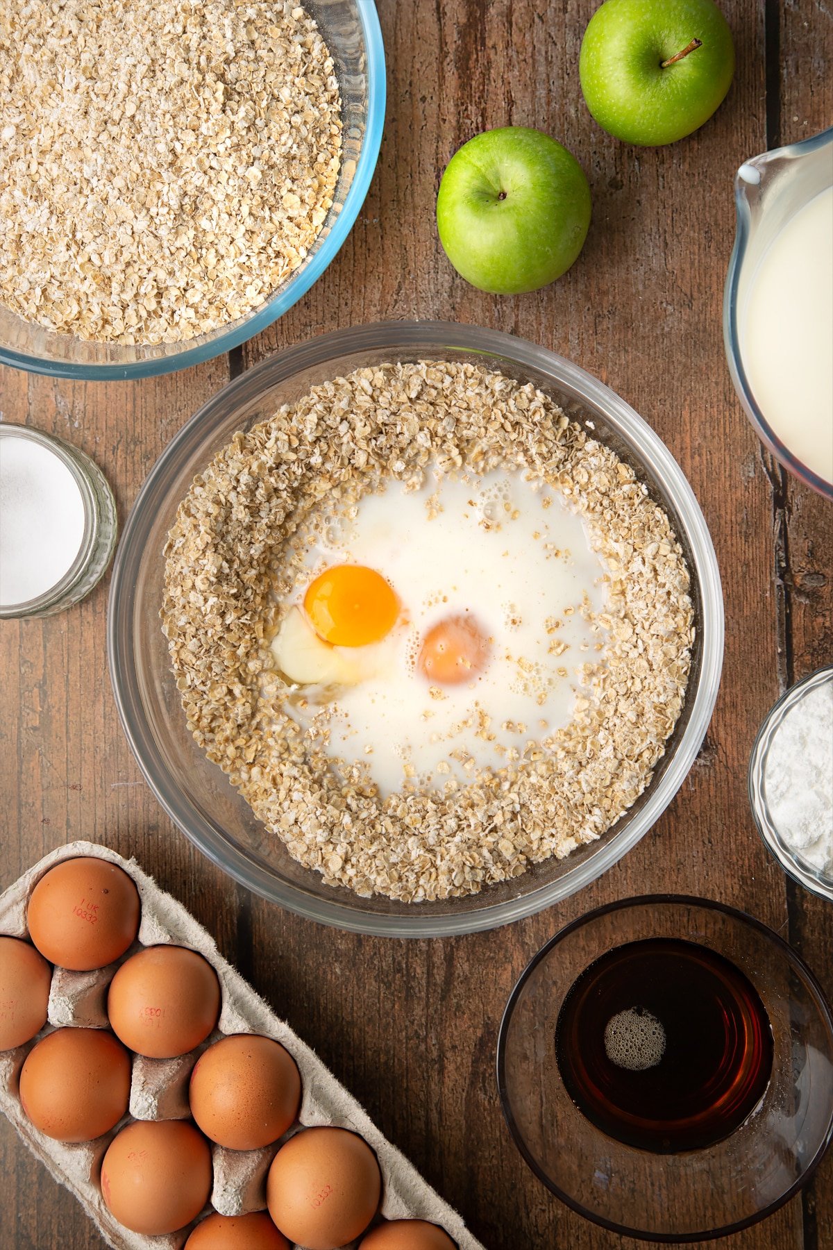 Oats, baking powder, salt, milk, eggs and maple syrup in a glass mixing bowl. Ingredients to make porridge squares surround the bowl.