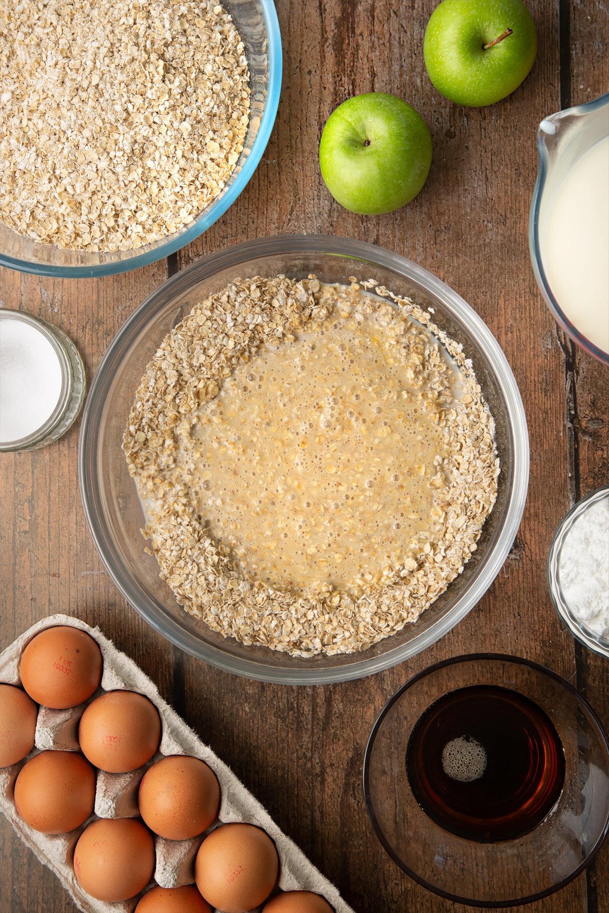 Oats, baking powder, salt, milk, eggs and maple syrup, semi mixed in a glass mixing bowl. Ingredients to make porridge squares surround the bowl.