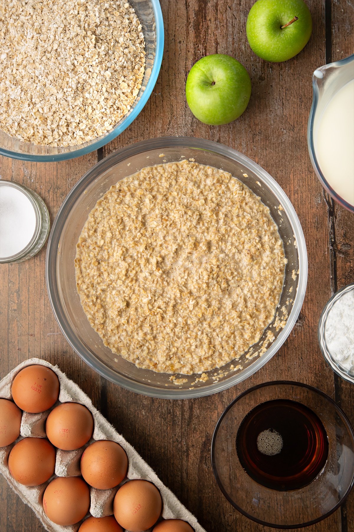 Oats, baking powder, salt, milk, eggs and maple syrup, mixed in a glass mixing bowl. Ingredients to make porridge squares surround the bowl.