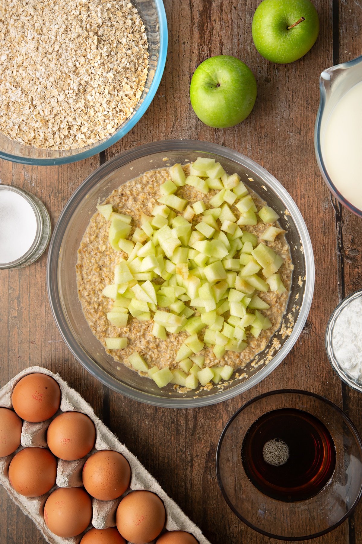 Oats, baking powder, salt, milk, eggs and maple syrup, mixed in a glass mixing bowl with apple pieces on top. Ingredients to make porridge squares surround the bowl.