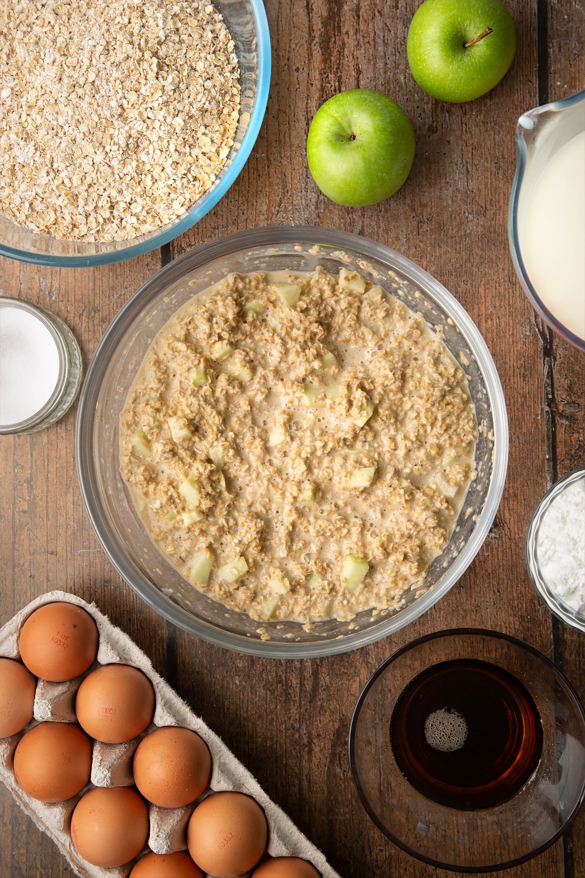 Oats, baking powder, salt, milk, eggs, maple syrup and apple mixed in a glass mixing bowl. Ingredients to make porridge squares surround the bowl.