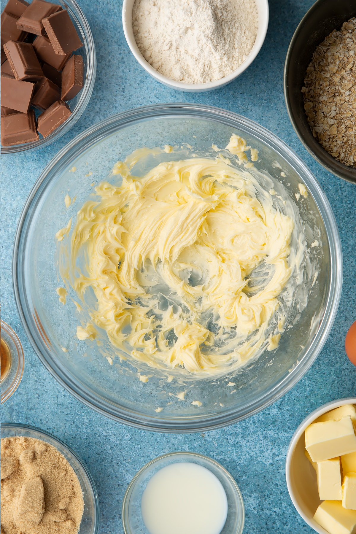 Whipped butter in a glass mixing bowl. Ingredients to make cookie monster cookies surround the bowl.