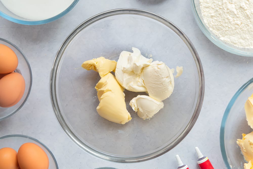 Margarine and cream cheese in a large glass mixing bowl. Ingredients to make filled red velvet cake surround the bowl. 