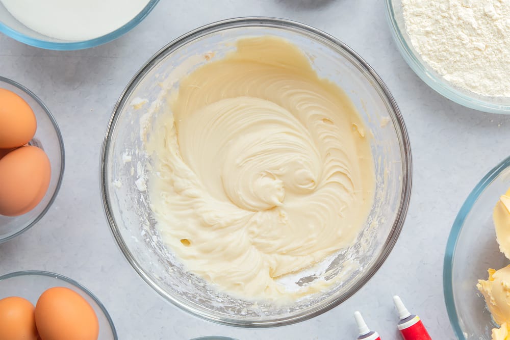 Margarine, cream cheese, icing sugar and vanilla extract whisked together in a large glass mixing bowl. Ingredients to make filled red velvet cake surround the bowl. 