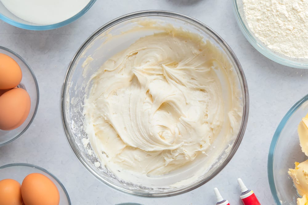 Cream cheese frosting in a large glass mixing bowl. Ingredients to make filled red velvet cake surround the bowl. 