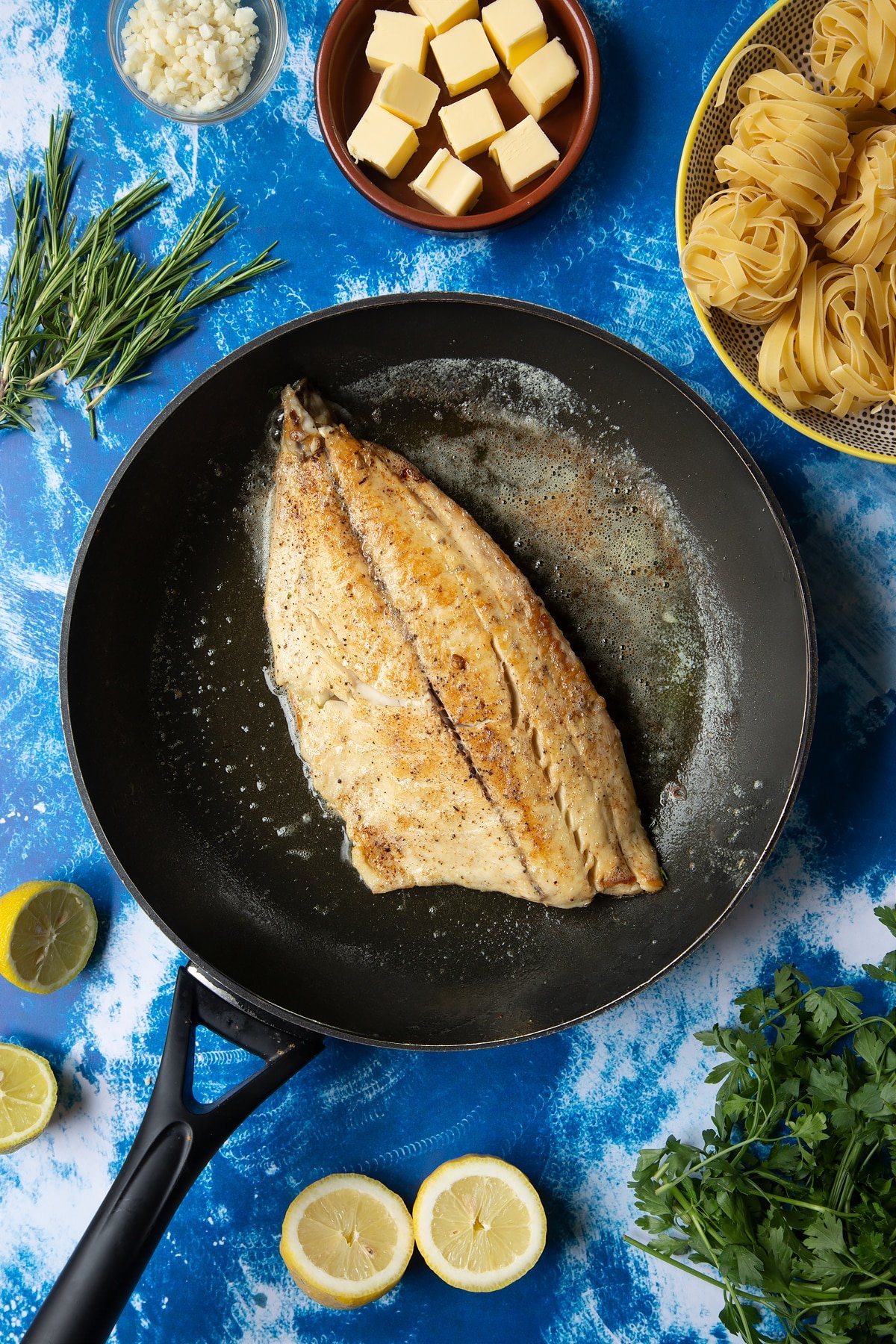 A large frying pan with melted butter and a fish fillet, skin side down. The fillet is cooked and lightly golden. Ingredients to make Dutch Yellowtail tagliatelle surround the pan.