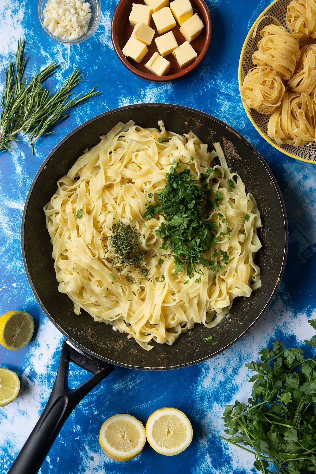 A large frying pan with tagliatelle topped with piles of finely chopped rosemary and parsley. Ingredients to make Dutch Yellowtail tagliatelle surround the pan.