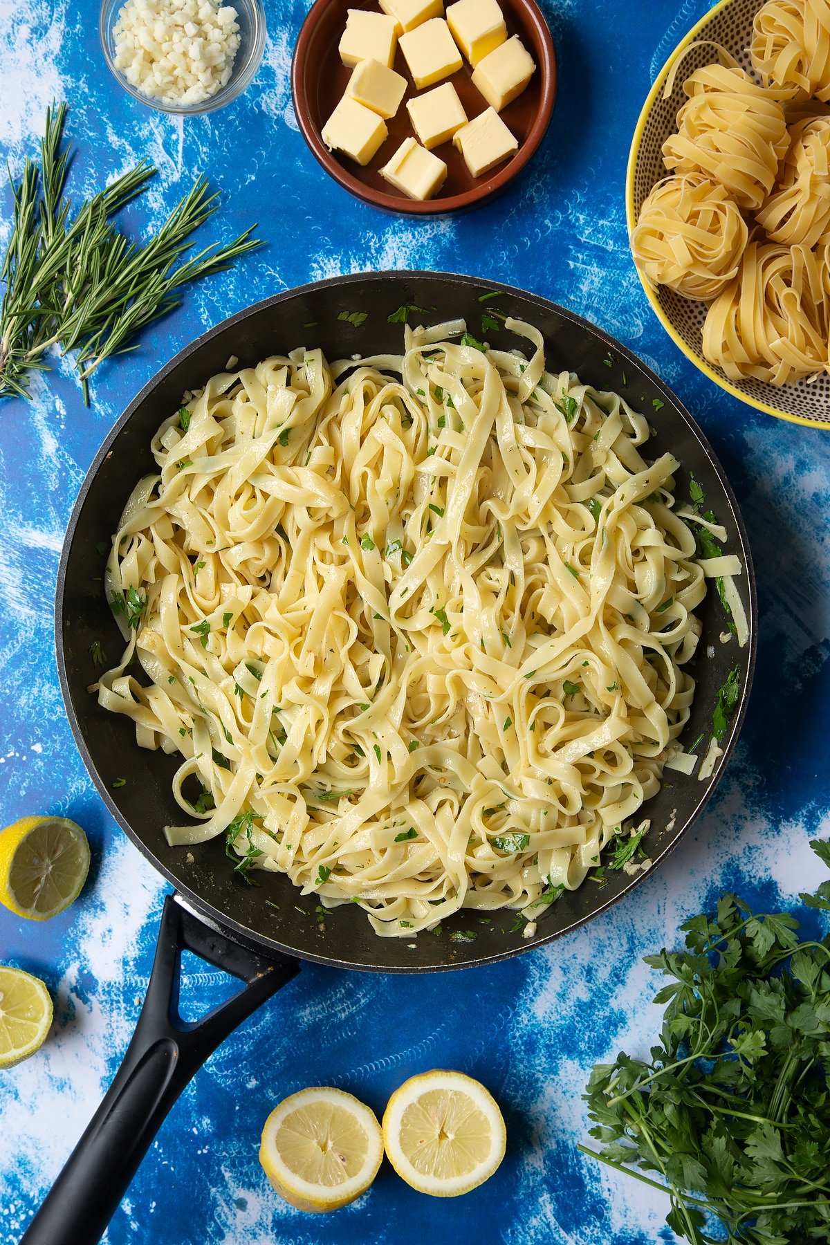 A large frying pan with tagliatelle, rosemary and parsley. Ingredients to make Dutch Yellowtail tagliatelle surround the pan.