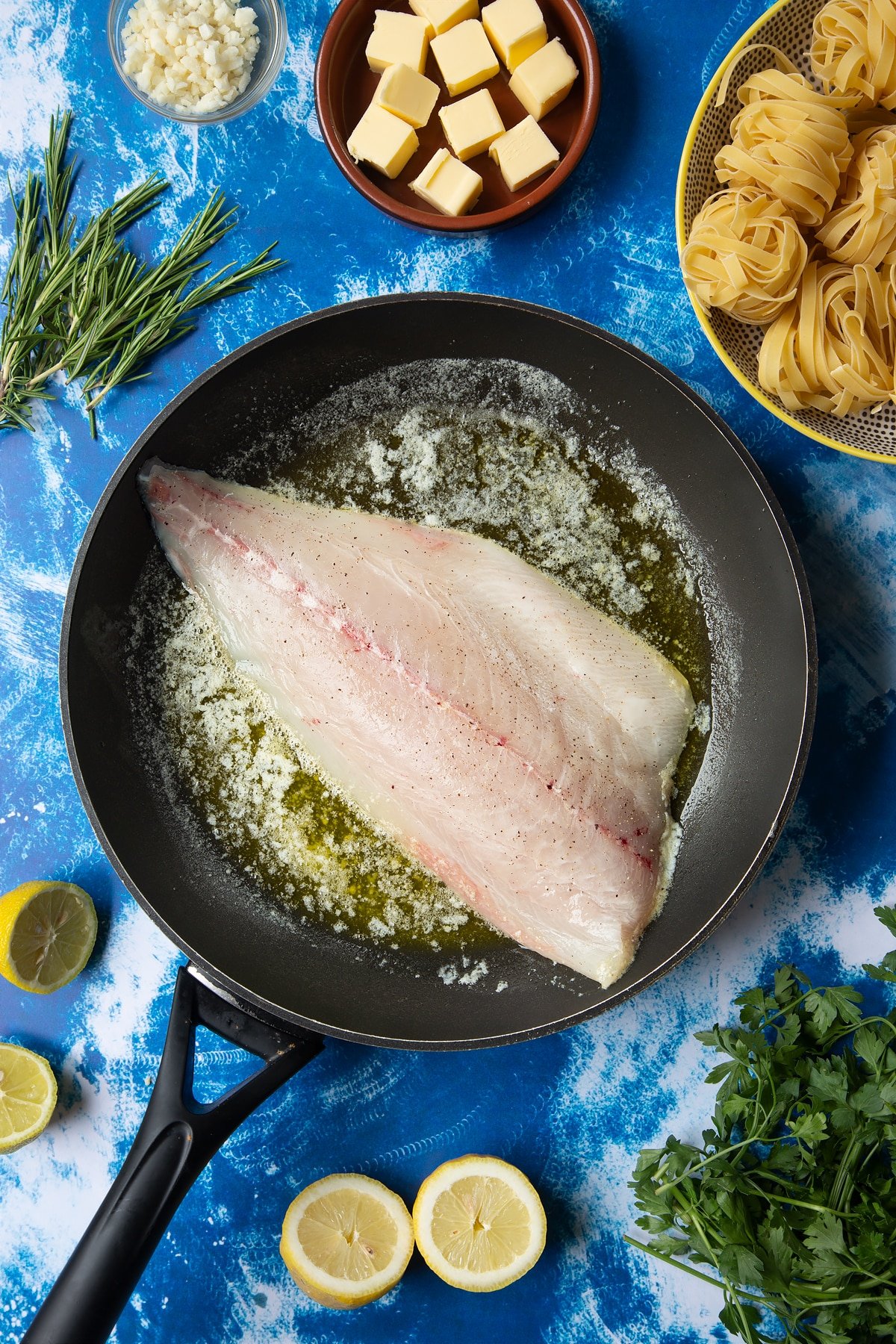 A large frying pan with melted butter and a fish fillet, skin side down. Ingredients to make Dutch Yellowtail tagliatelle surround the pan.