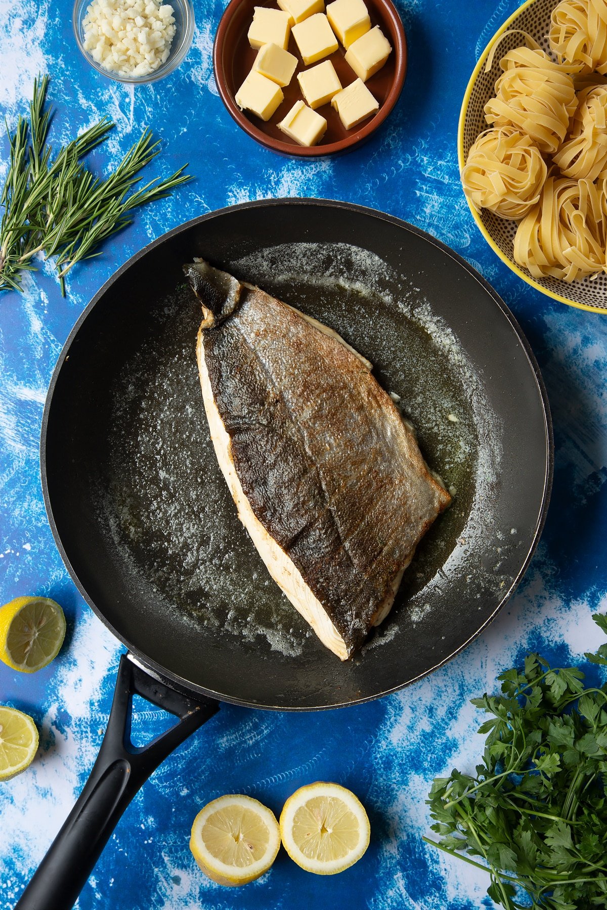 A large frying pan with melted butter and a fish fillet, skin side up. The skin is cooked and crisp. Ingredients to make Dutch Yellowtail tagliatelle surround the pan.