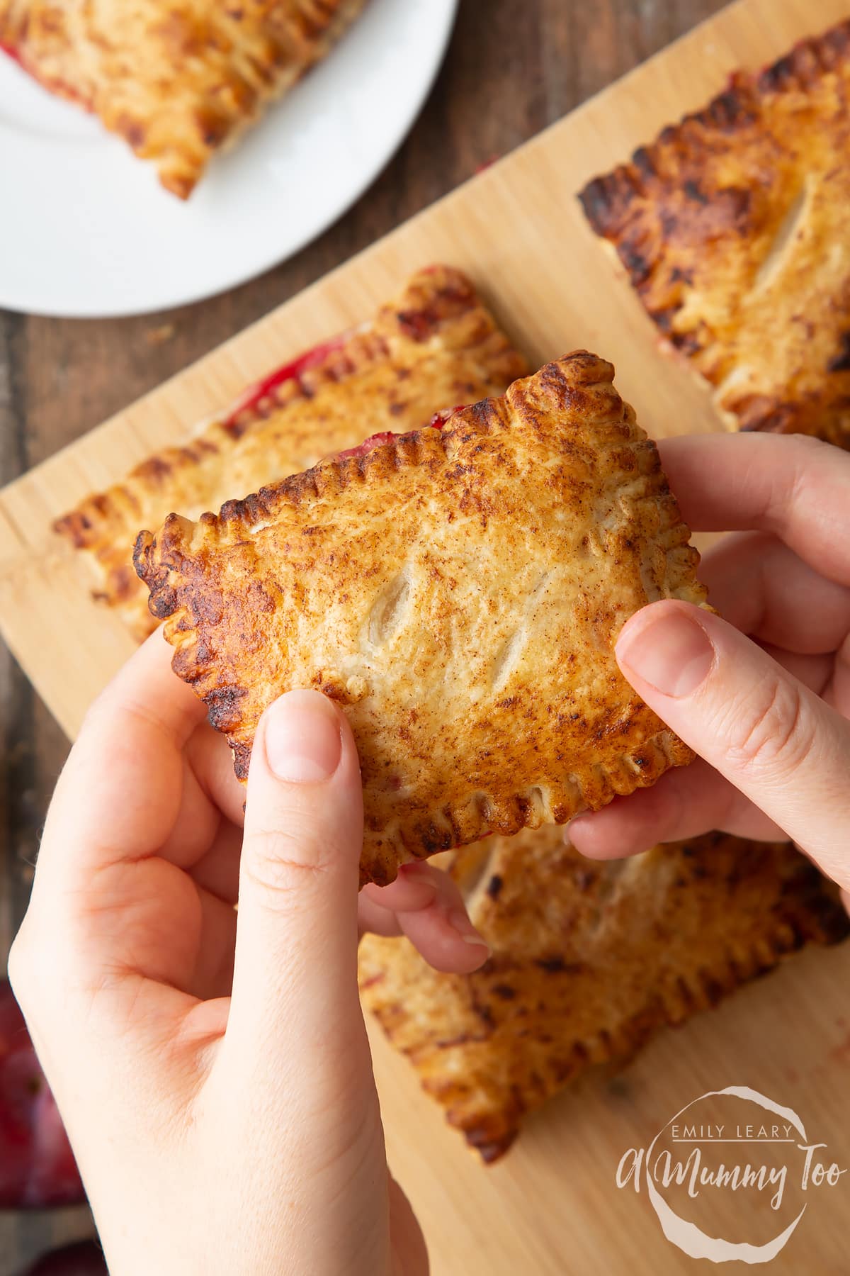 Two hands holding a plum pastry. More pastries are shown on a board in the background.