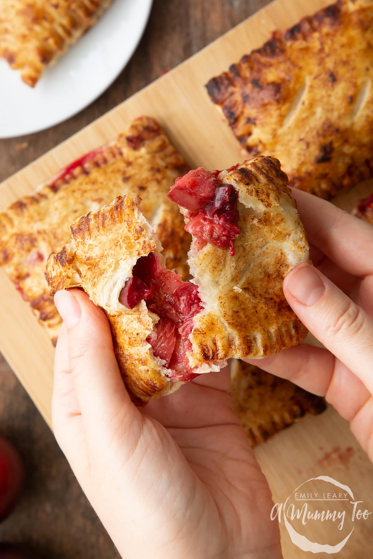 Two hands holding a plum pastry. It is broke open, showing the filling inside. More pastries are shown on a board in the background.