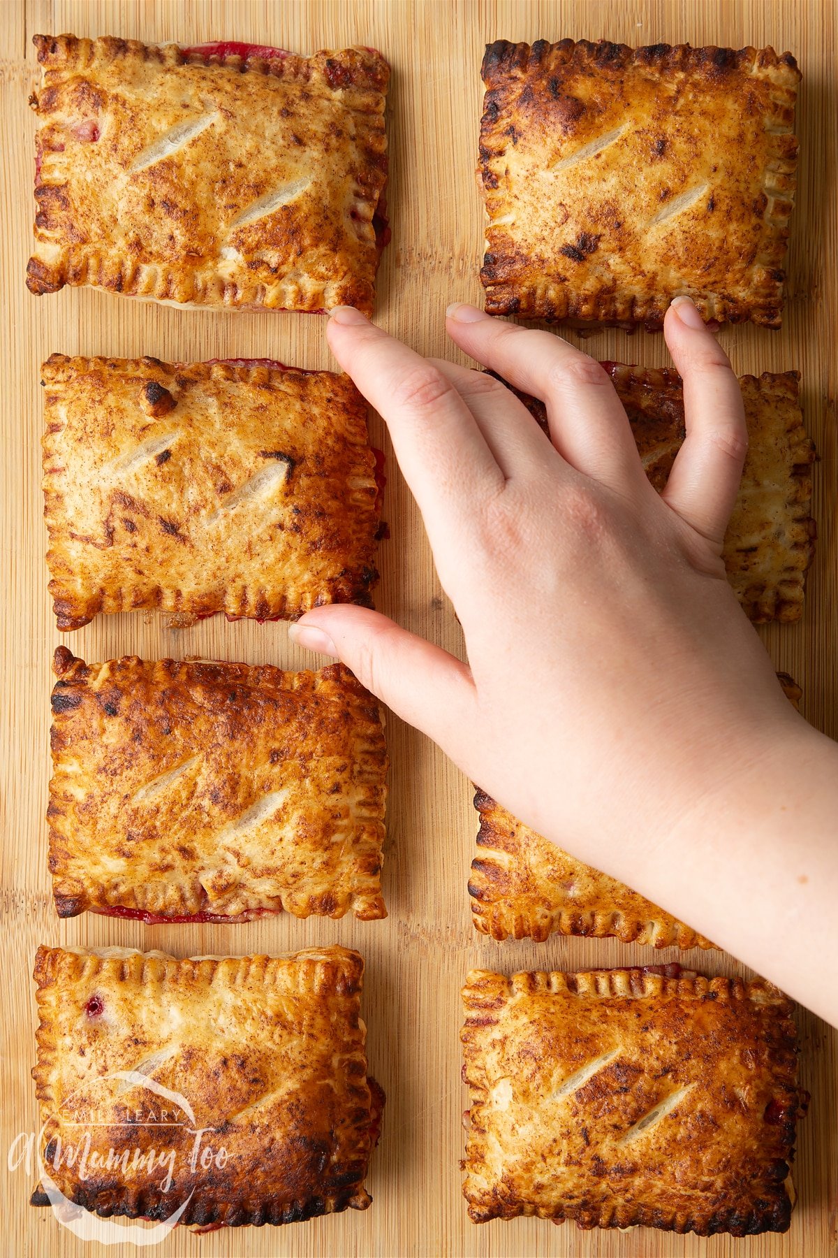 Eight plum pastries on a wooden board. A hand reaches to take one.