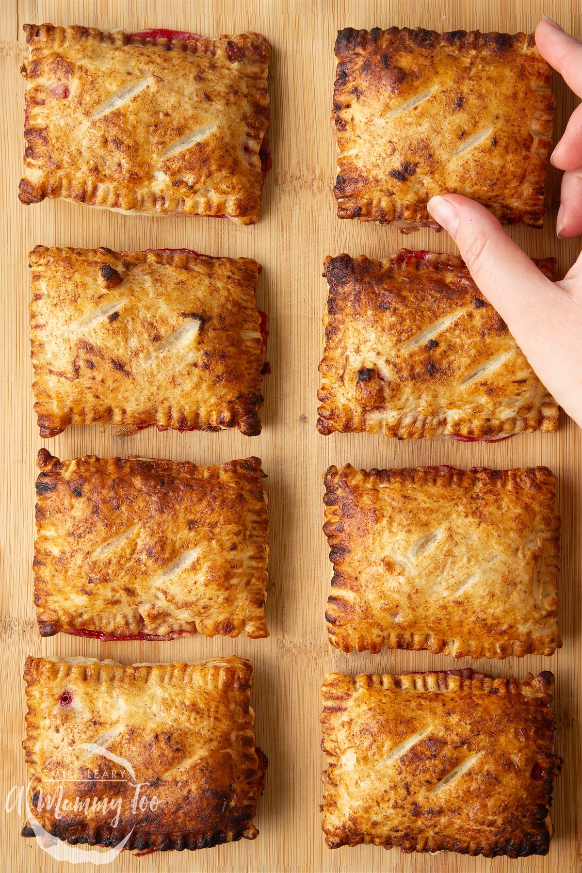 Eight plum pastries on a wooden board. A hand reaches to take the one in the top corner.