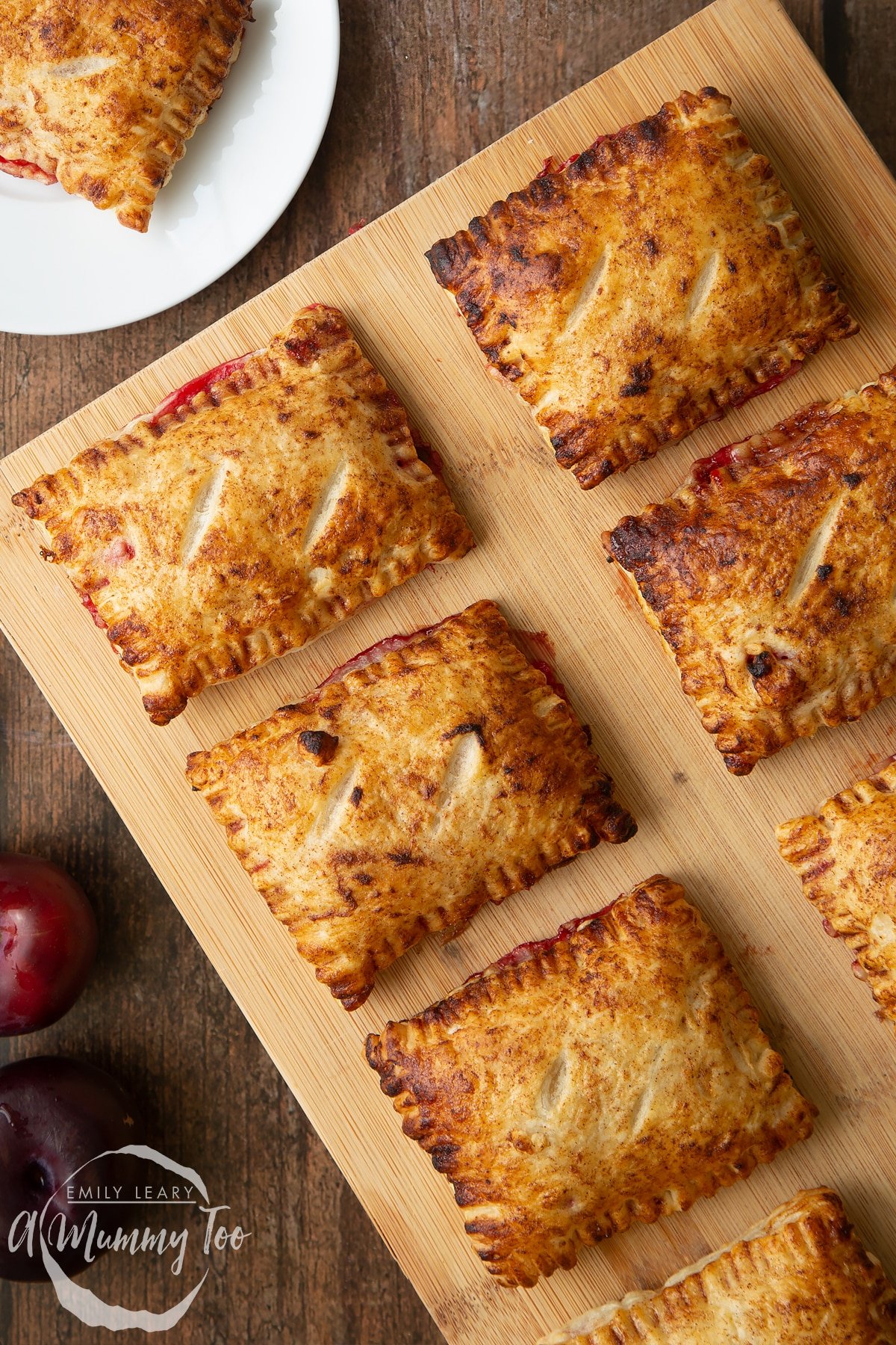 Plum pastries on a wooden board. To the edges of frame, a plum pastry is shown on white plate.