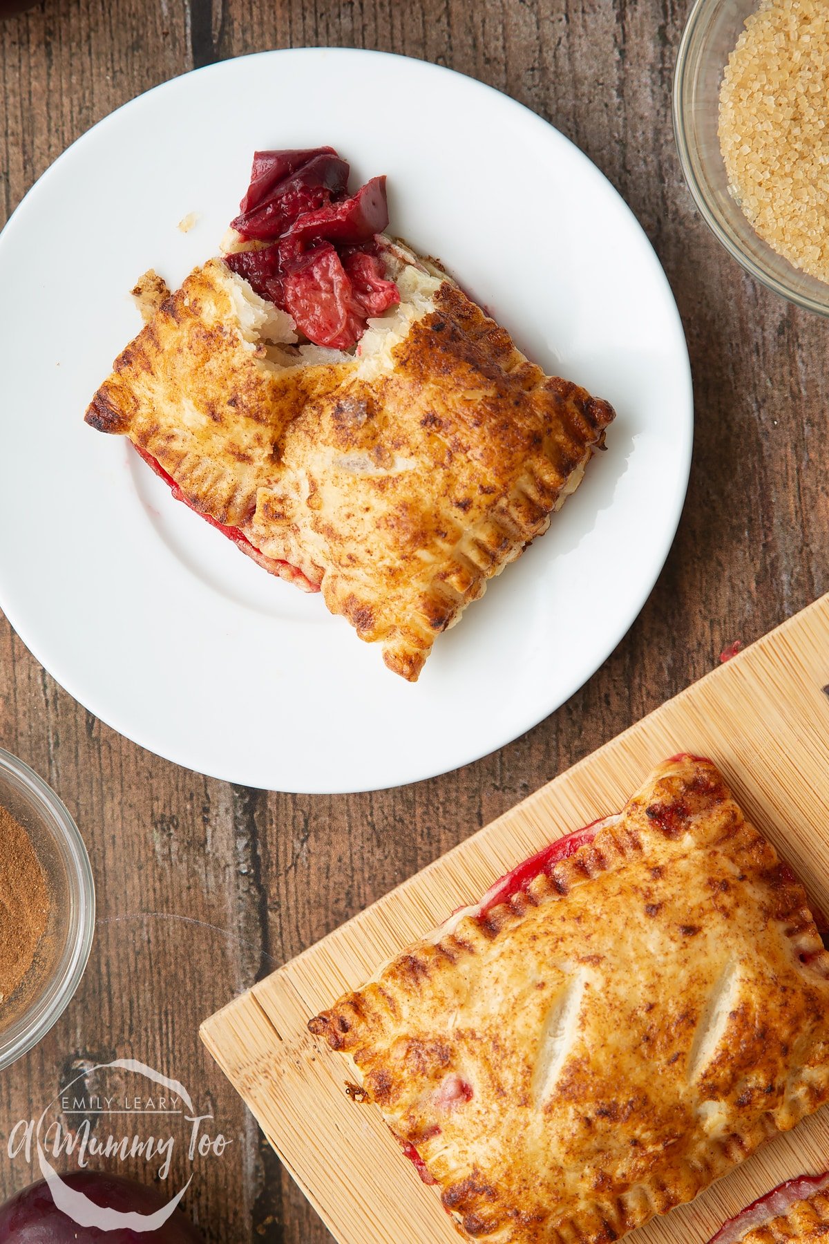 A plum pastry on white plate. The corner is broken open with the filling showing. Another plum pastry is shown on a wooden board.