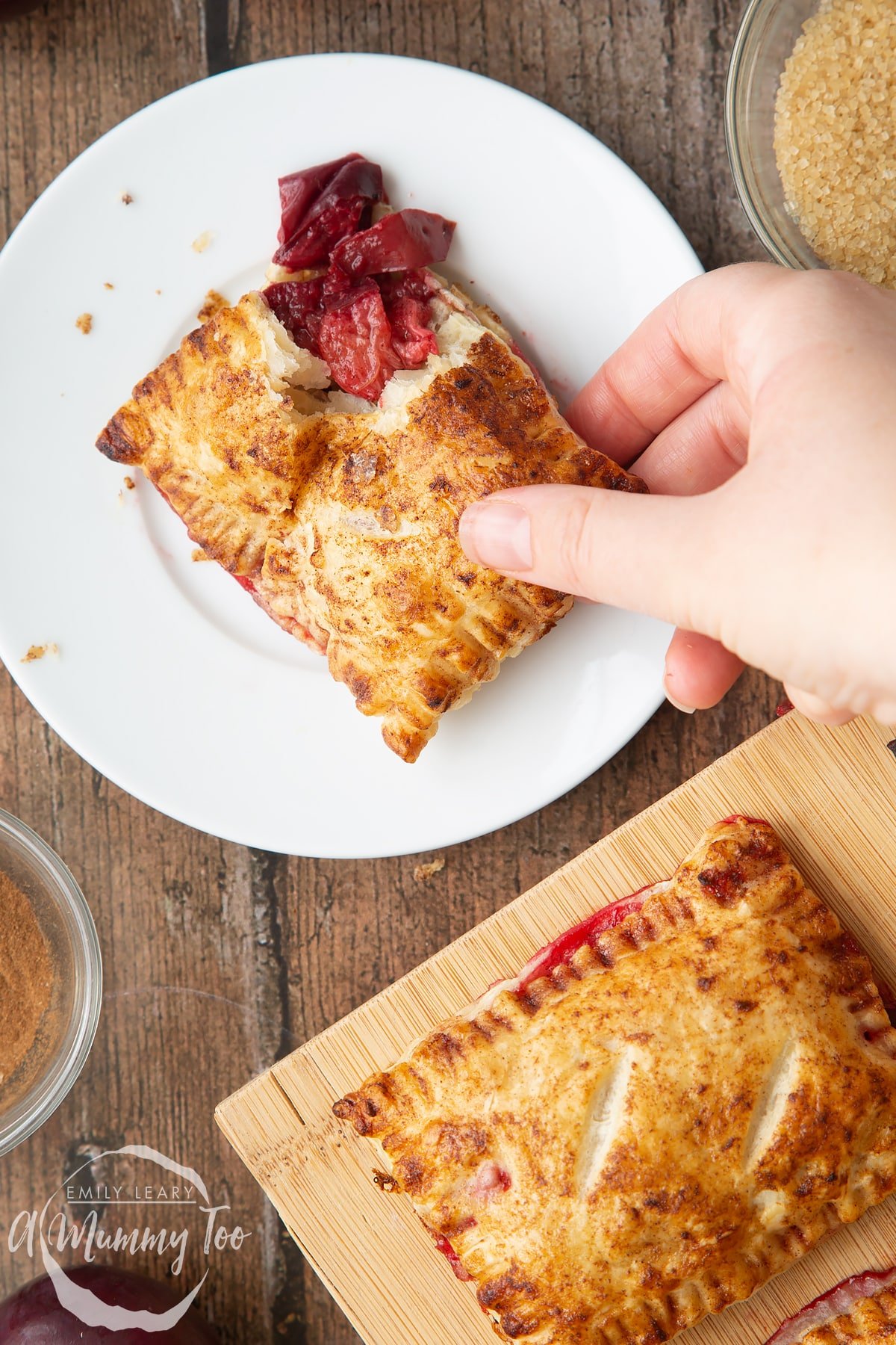 A plum pastry on white plate. The corner is broken open with the filling showing. A hand reaches to take it. Another plum pastry is shown on a wooden board.