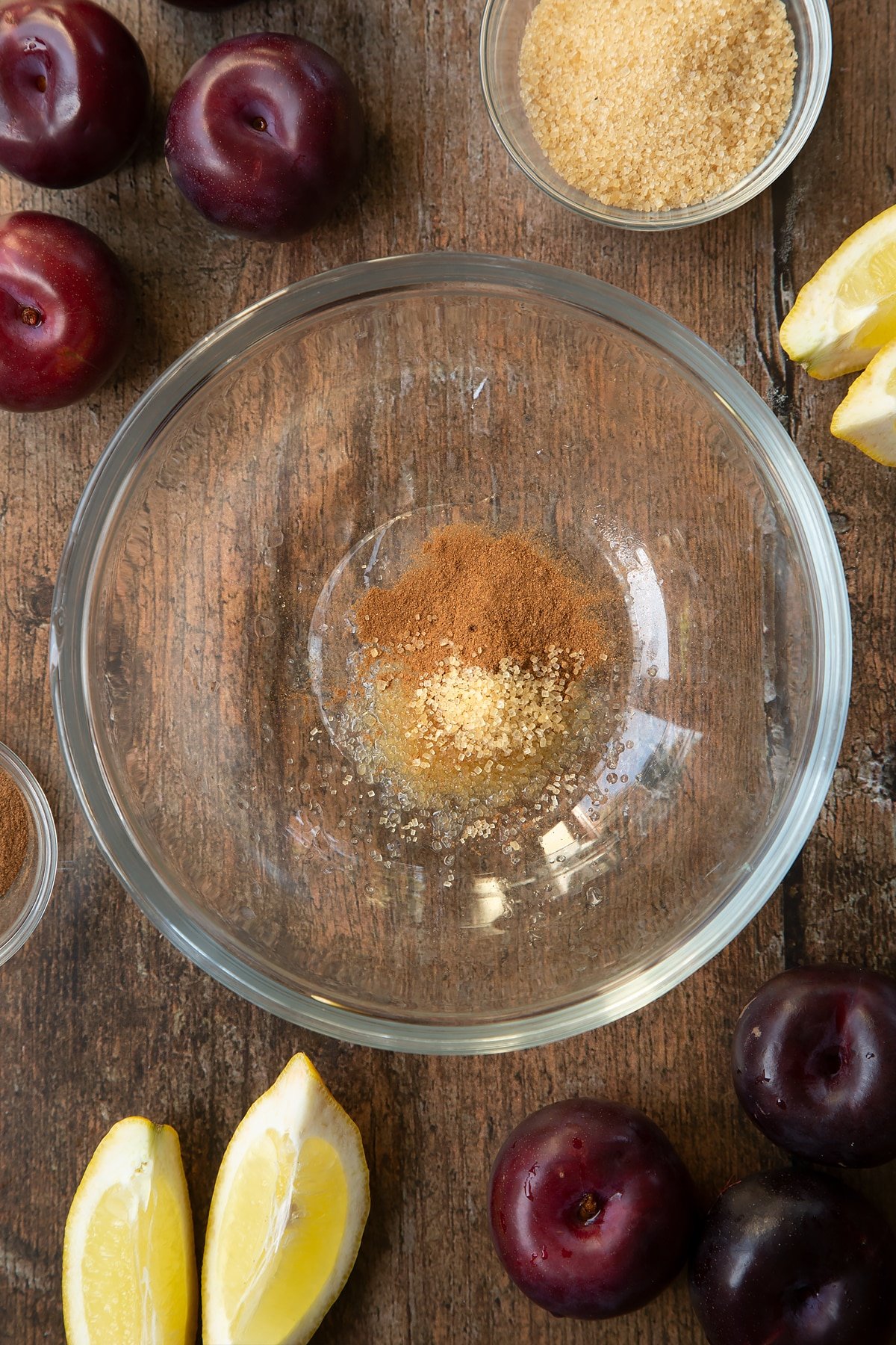 Sugar, lemon juice and cinnamon in a glass mixing bowl. Ingredients to make a plum pastry recipe surround the bowl.
