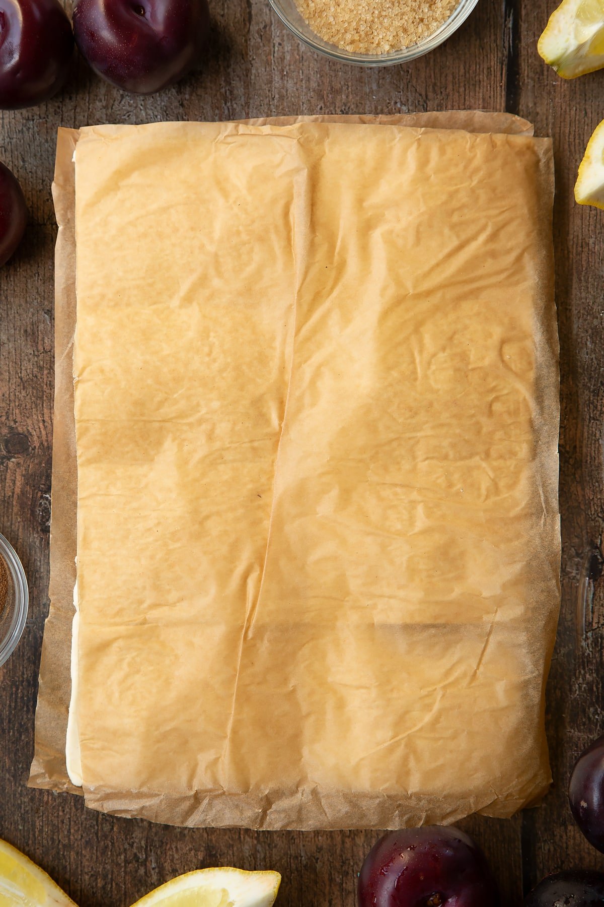 A puff pastry sheet topped with chopped plums and another sheet laying on top of it covered with paper. Ingredients to make a plum pastry recipe surround the pastry.