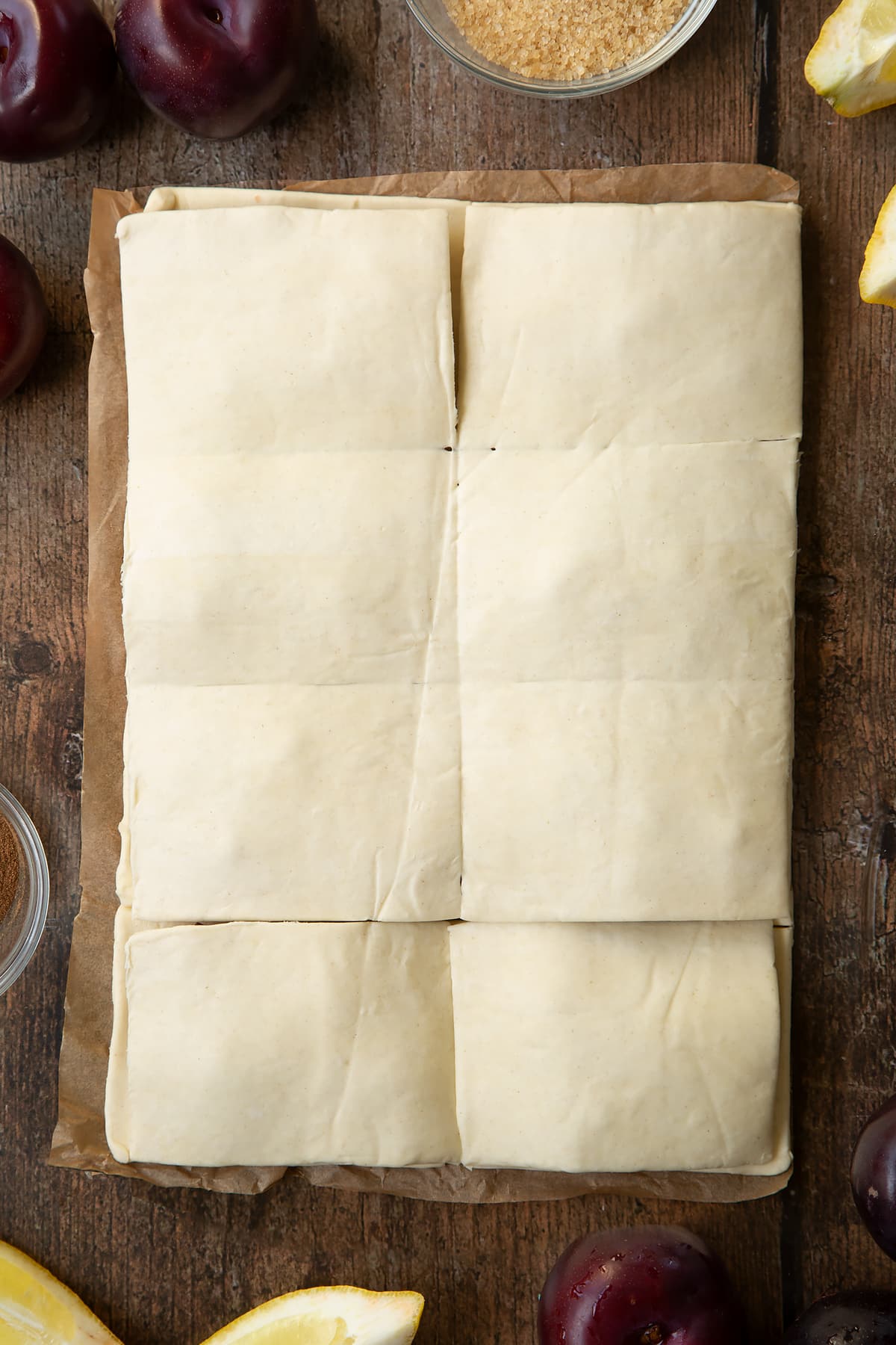 A puff pastry sheet topped with chopped plums and another sheet laying on top of it. Ingredients to make a plum pastry recipe surround the pastry.