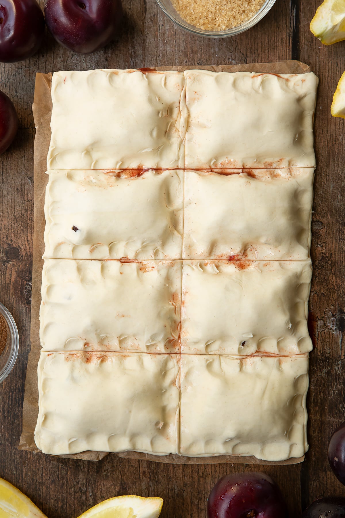 Puff pastry parcels with the edges pressed with finger tips. Ingredients to make a plum pastry recipe surround the pastry.