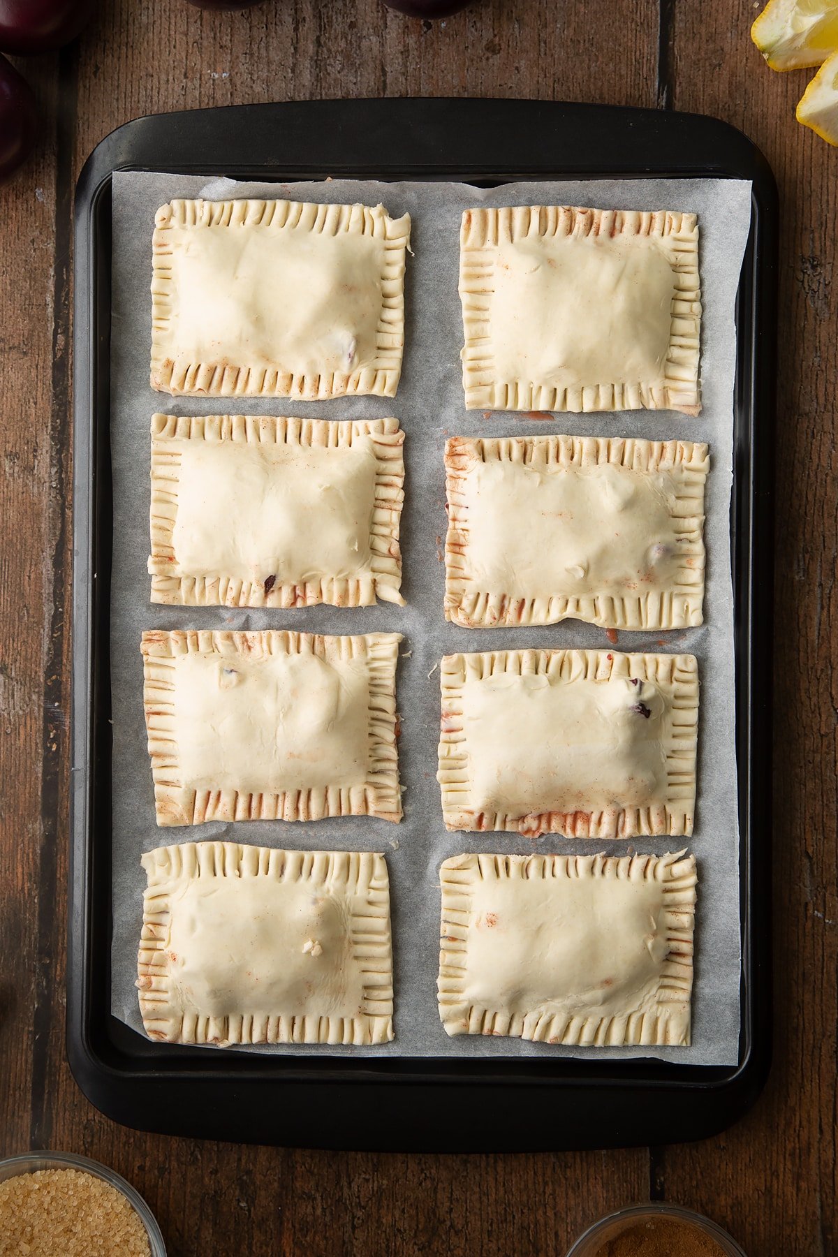 Puff pastry parcels on a baking paper lined tray. The edges are crimped with a fork and trimmed. Ingredients to make a plum pastry recipe surround the tray.