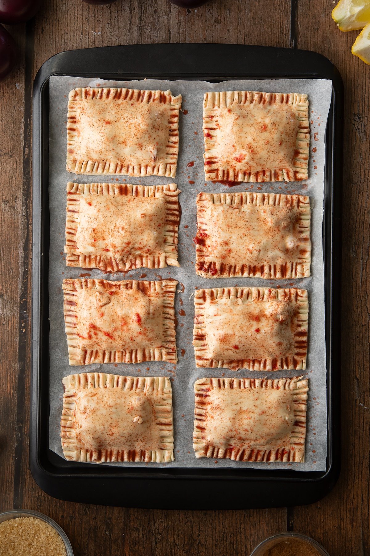 Puff pastry parcels on a baking paper lined tray. The parcels have been brush with plum juice. Ingredients to make a plum pastry recipe surround the tray.