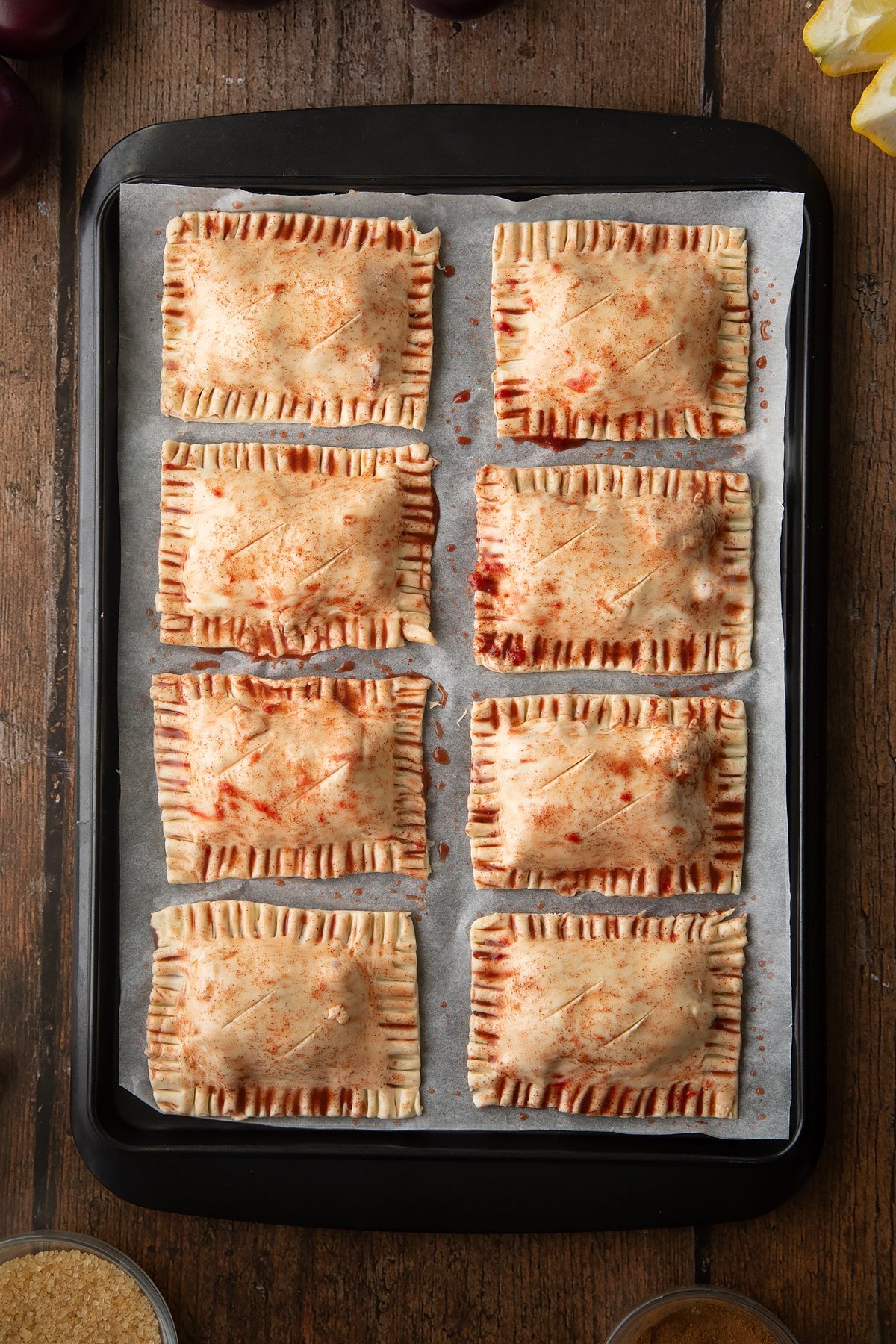 Puff pastry parcels on a baking paper lined tray. The parcels have been brush with plum juice. Slits have been cut across the top of each. Ingredients to make a plum pastry recipe surround the tray.