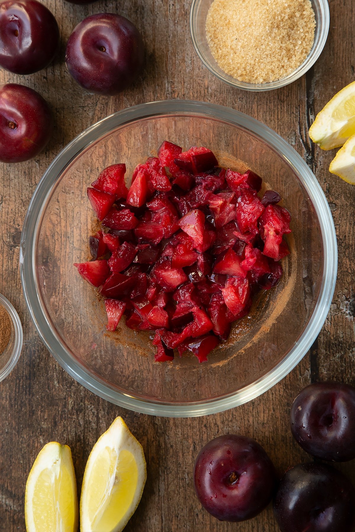 Sugar, lemon juice, cinnamon and plums in a glass mixing bowl. Ingredients to make a plum pastry recipe surround the bowl.