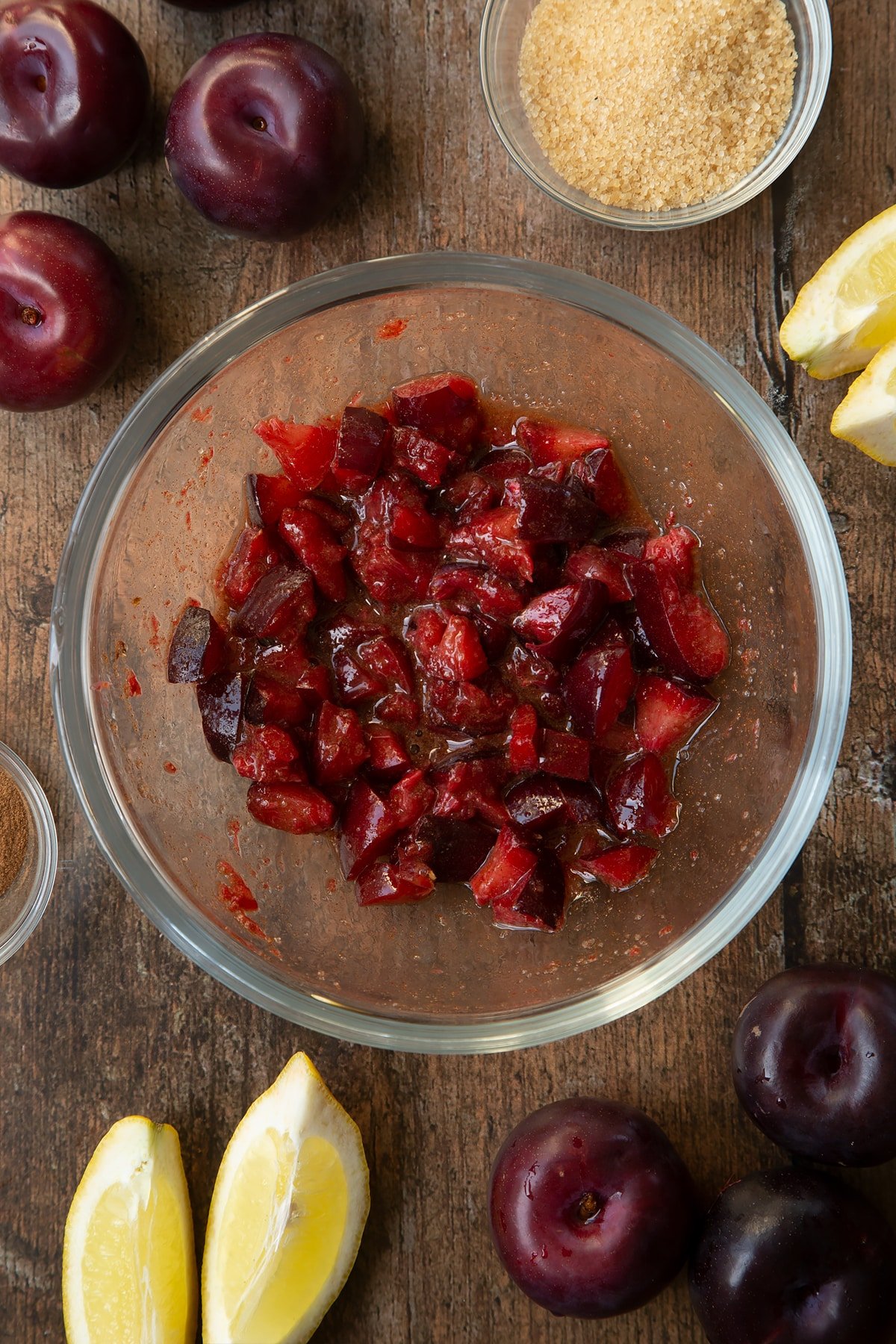 Sugar, lemon juice, cinnamon and plums mixed together in a glass mixing bowl. Ingredients to make a plum pastry recipe surround the bowl.