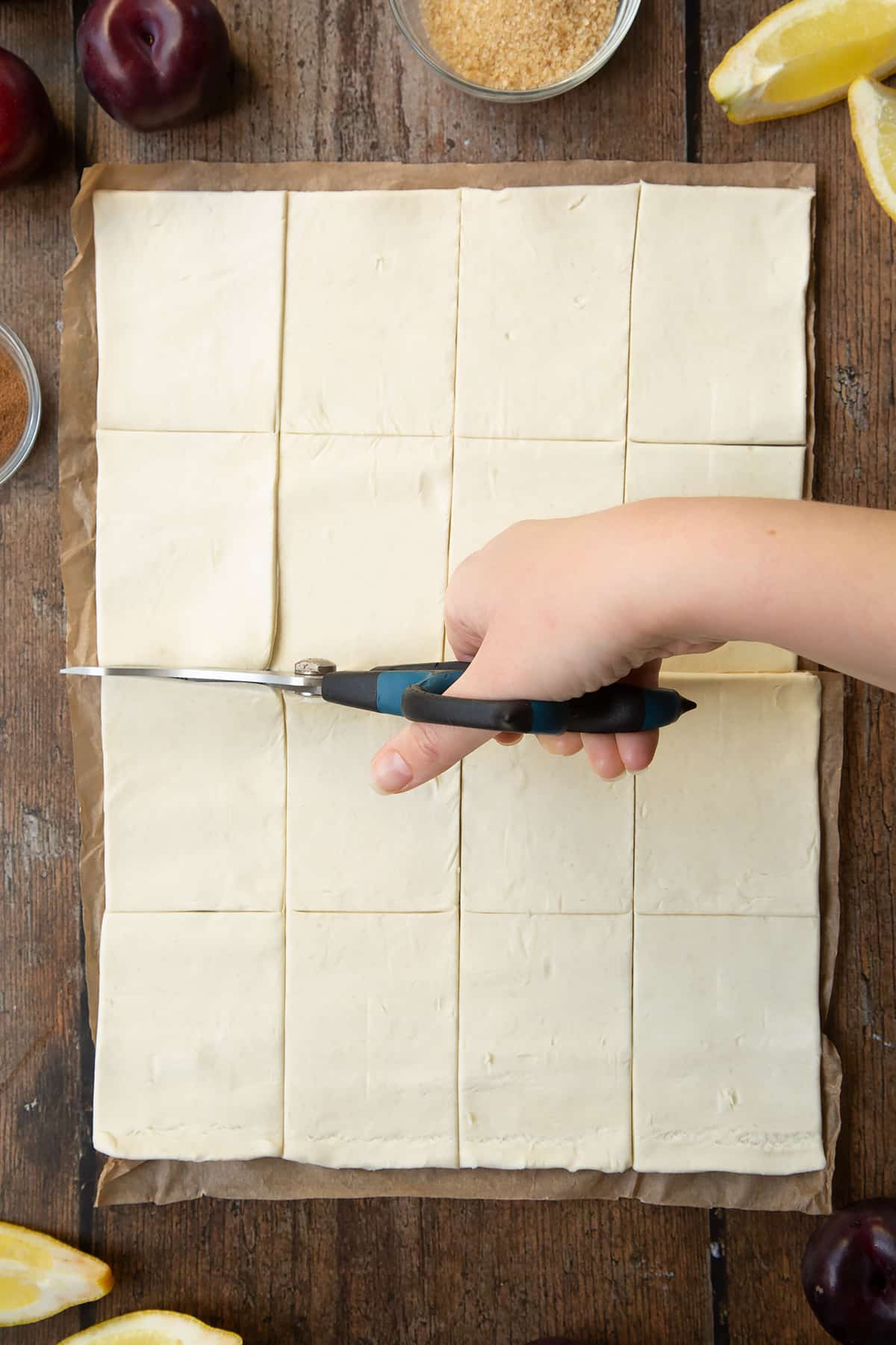 A puff pastry sheet cut into 16 pieces. Scissors are cutting the sheet in half. Ingredients to make a plum pastry recipe surround the pastry.