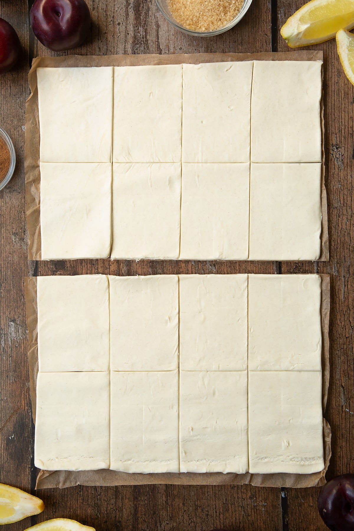 A puff pastry sheet cut into 16 pieces. The sheet is cut in half. Ingredients to make a plum pastry recipe surround the pastry.