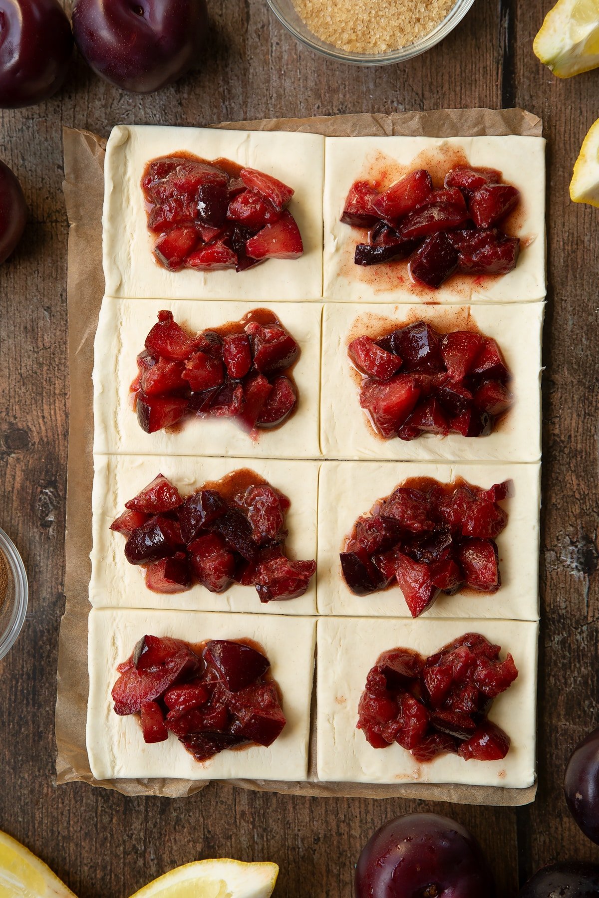 A puff pastry sheet cut into 8 pieces. Each piece is topped with chopped plums. Ingredients to make a plum pastry recipe surround the pastry.